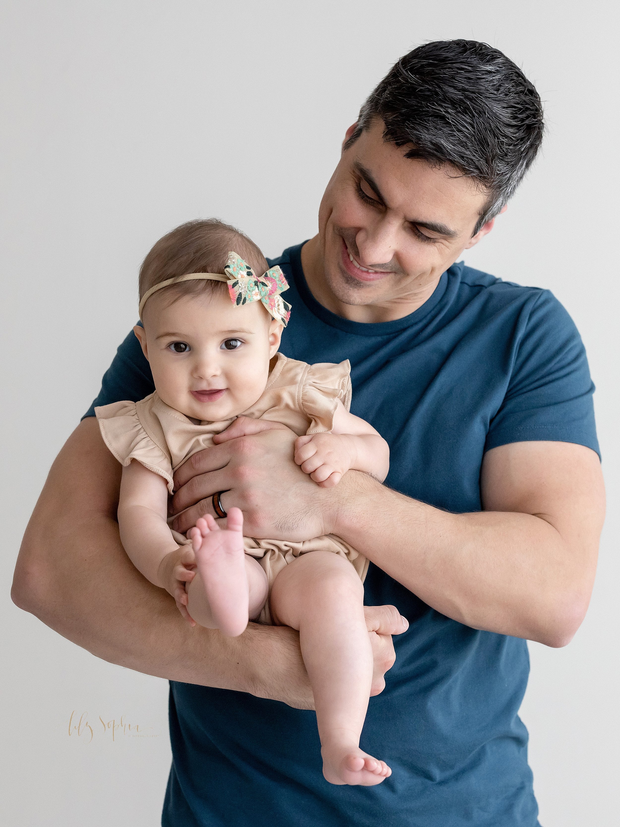  Milestone picture of a father holding his one year old daughter in front of him as she sticks up her right foot taken near Virginia Highlands in Atlanta in a studio that uses natural light. 