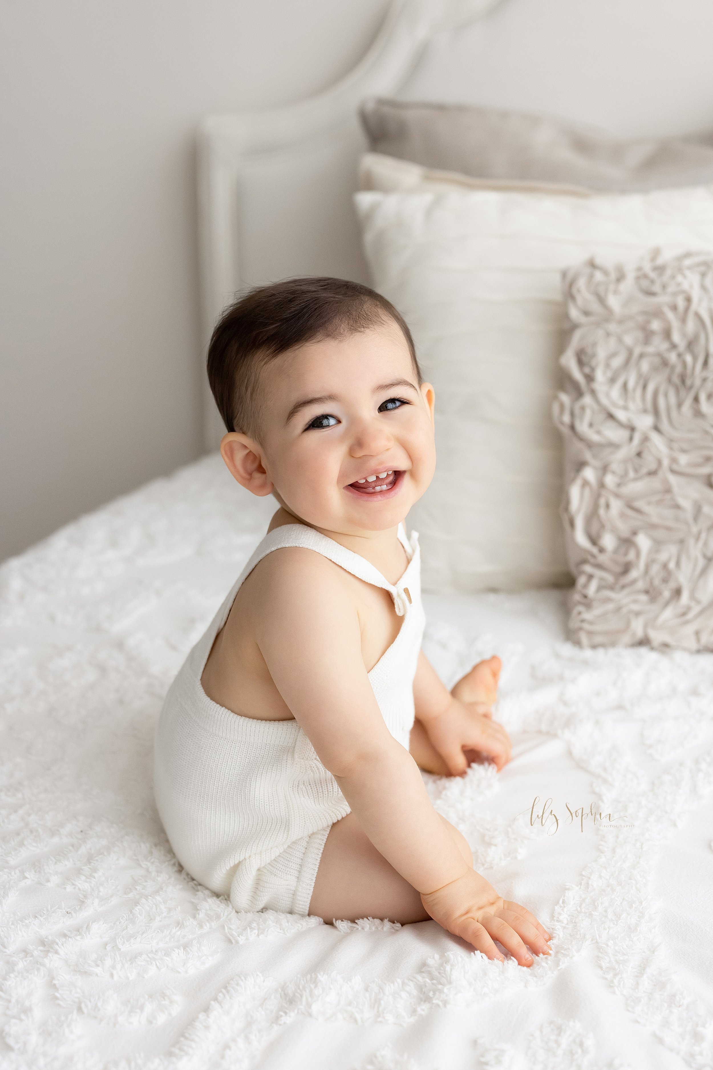  First birthday photo session of a smiling one year old boy as he sits atop a bed and looks over his right shoulder taken near Brookhaven in Atlanta in a natural light studio. 