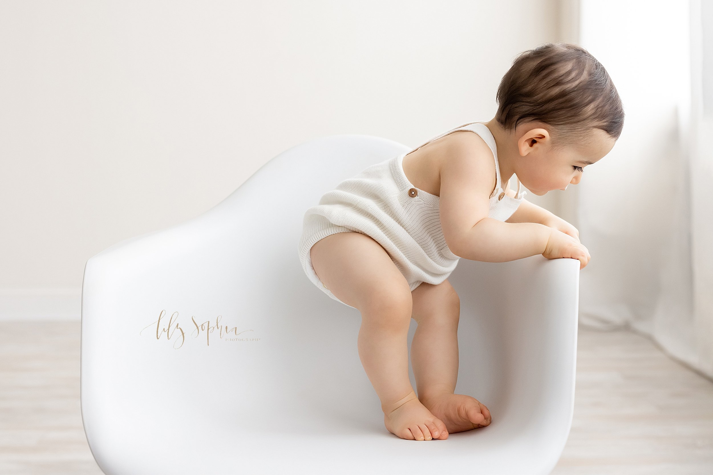  First birthday photo session of a curious one year old boy as he stands in a white molded chair holding onto the side and peers over the edge taken in front of a window streaming natural light in a photography studio near Buckhead in Atlanta. 