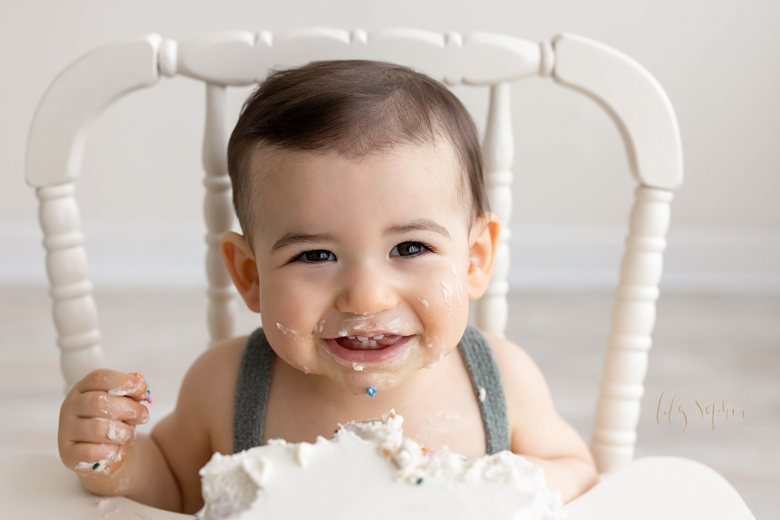  First birthday photo shoot with a one year old baby boy as he sits in an antique high chair smiling to show his teeth with icing from his smash cake on his face and his hand taken in a natural light studio near Ansley Park near Atlanta, Georgia. 