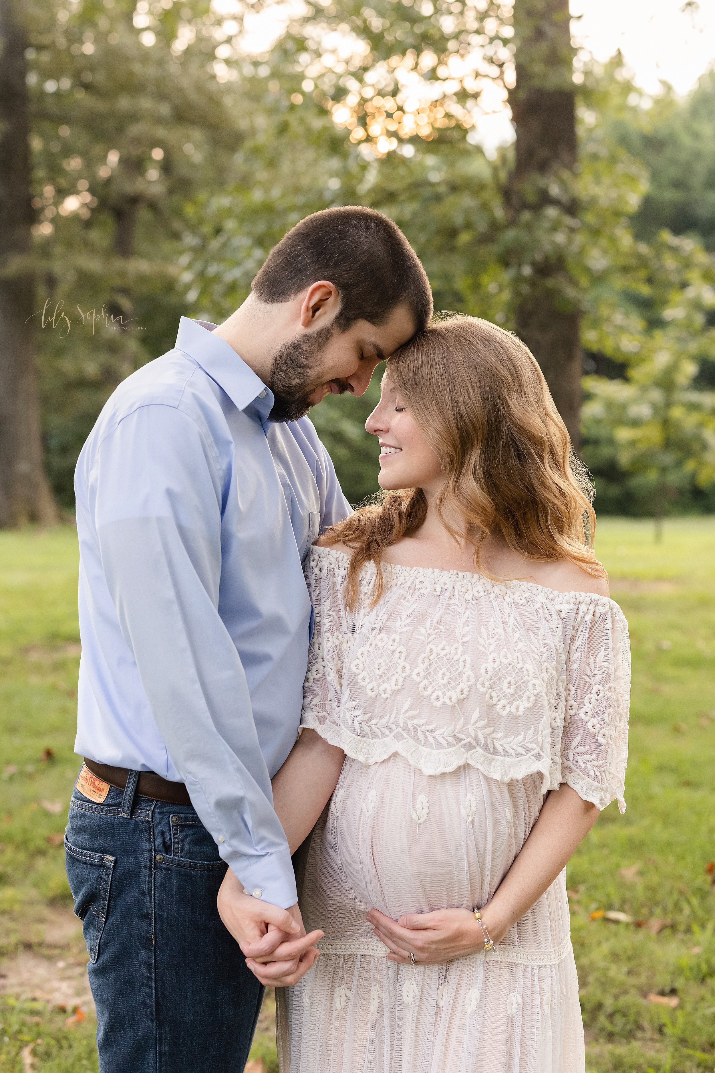  Maternity portrait of a pregnant mother  wearing an off the shoulder eyelet bodice gown as she turns her head over her left shoulder to look toward her husband who is standing at her side as he places his forehead on the top his wife’s head and the 