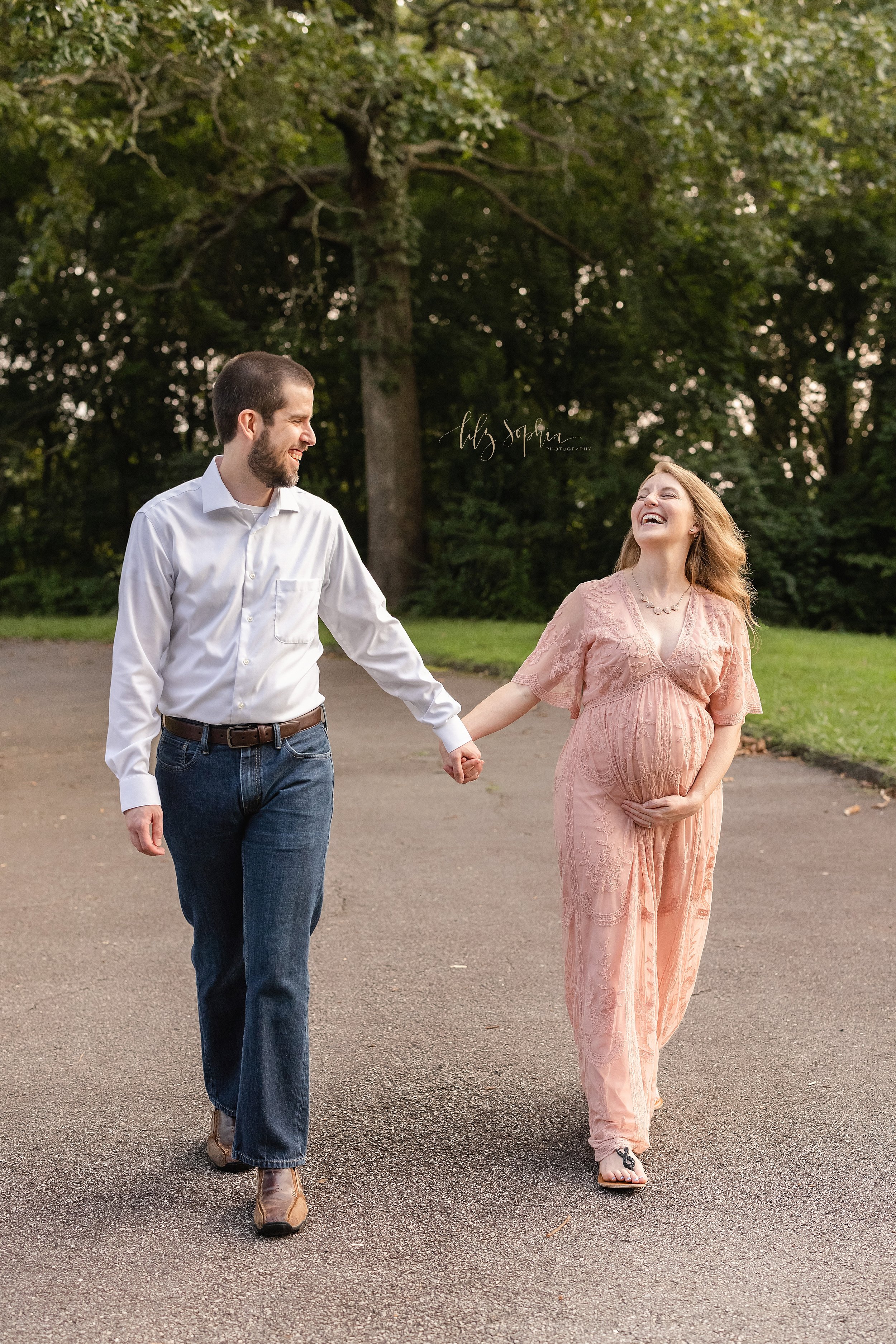  Maternity photo session of an expectant couple as they walk hand in hand down a path in an Atlanta park at sunset and laugh together. 