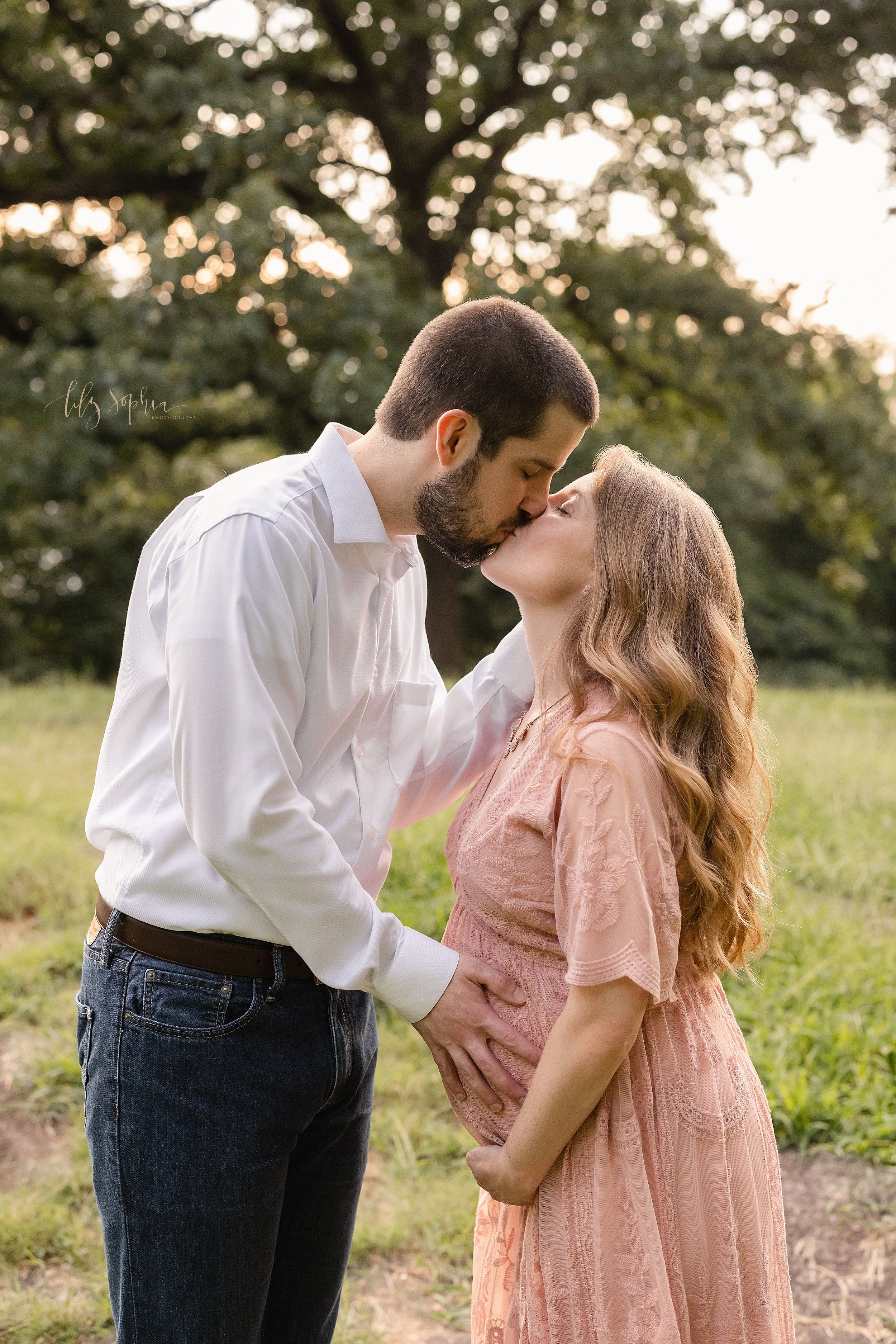  Maternity photo session on a mountain near Atlanta of a pregnant mother standing and holding the base of her belly as her husband faces her with his right hand on their child in utero and his left hand on her head as the two embrace taken at sunset.