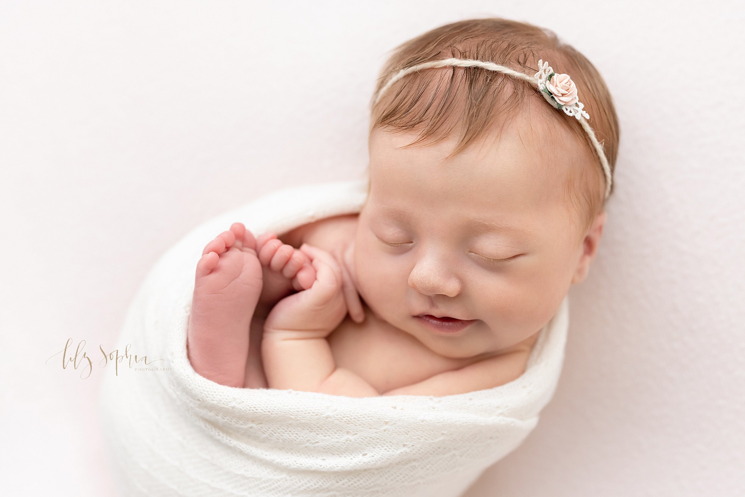  Newborn portrait of a peacefully sleeping infant girl who is nestled in a stretchy swaddle wearing a rose bud headband in her hair taken in a studio near Buckhead in Atlanta that uses natural light. 