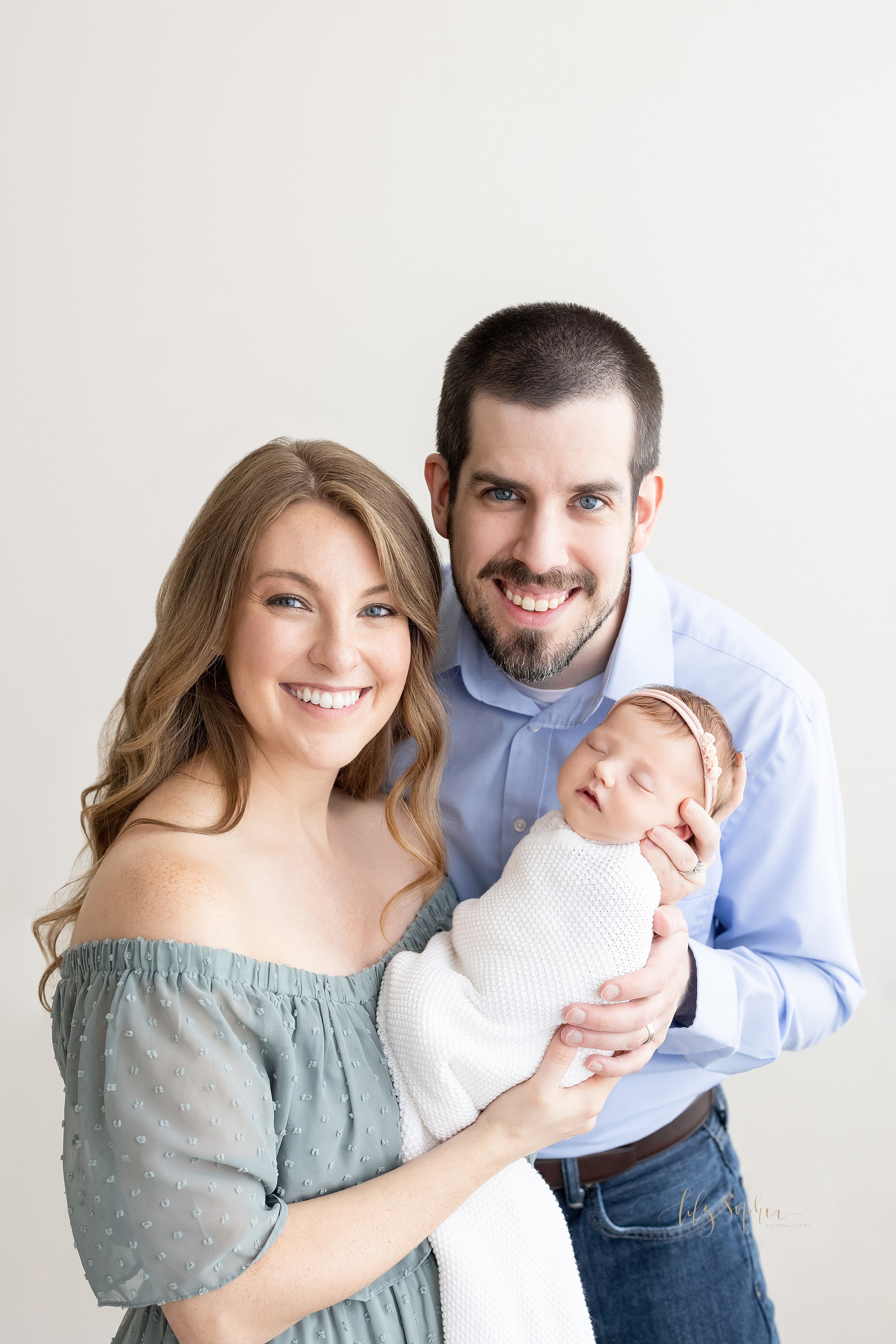  A happy family portrait celebrating their newborn baby with mom holding her infant daughter in her hands as dad stand behind her and places his hand on his daughter’s side taken in natural light in a photography studio near Oakhurst in Atlanta, Geog