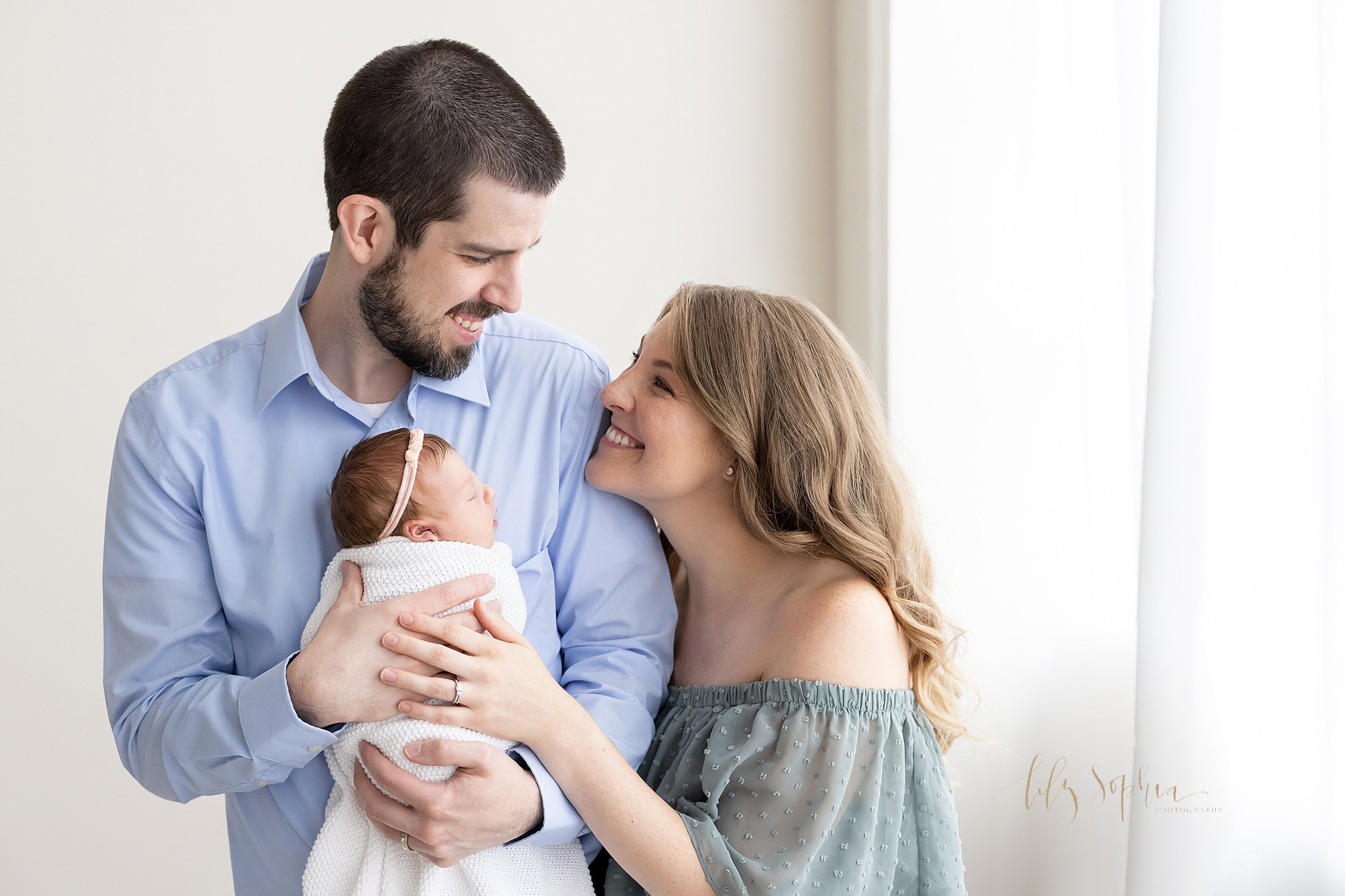  Newborn family picture of a father holding his infant daughter against his chest as mom stand to the left side of her husband and places her hand on top her husband’s as the couple stand and smile proudly at one another in front of a window streamin