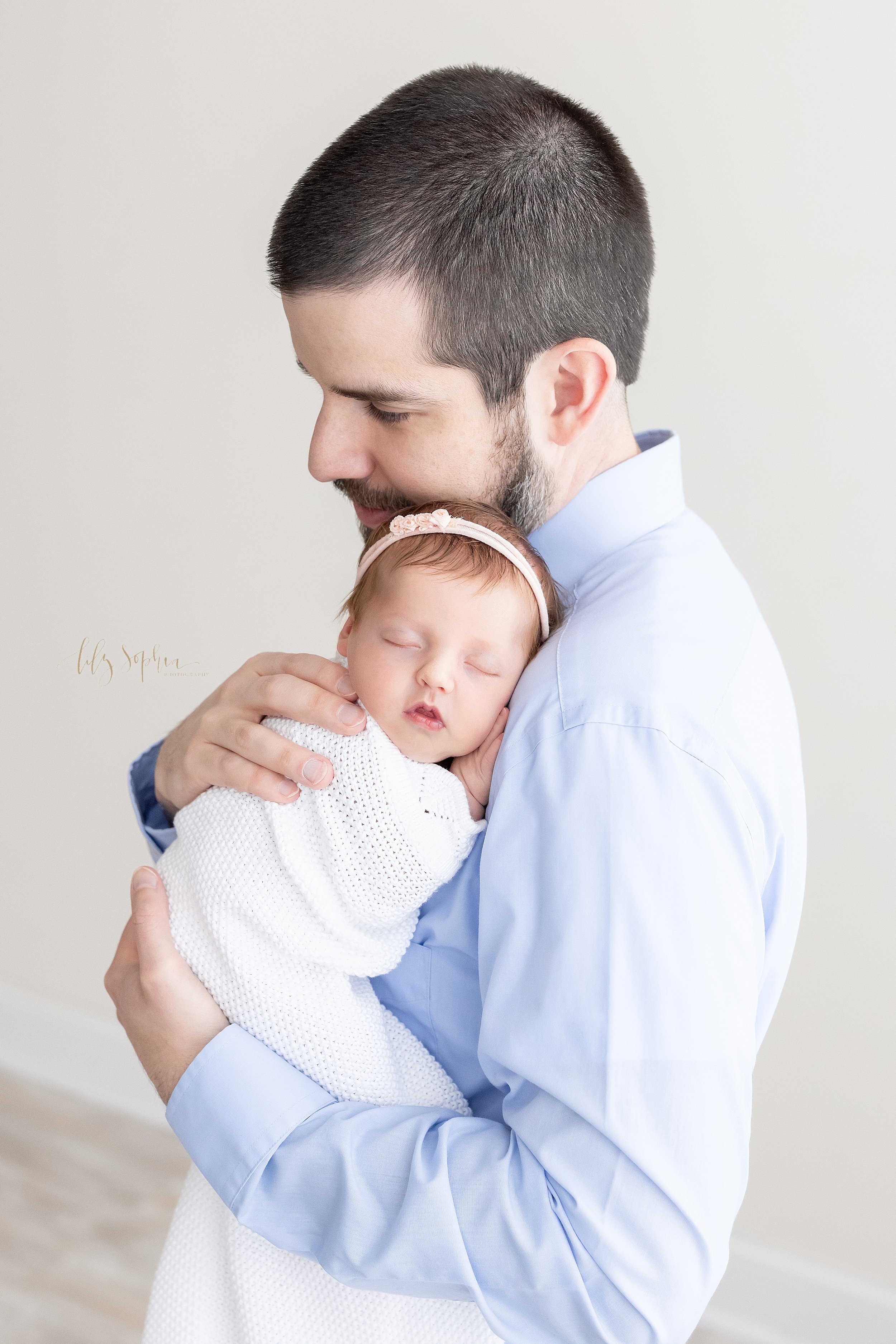  Newborn portrait of a father holding his peacefully sleeping infant daughter against his chest as he stands in a natural light studio near Midtown in Atlanta, Georgia. 