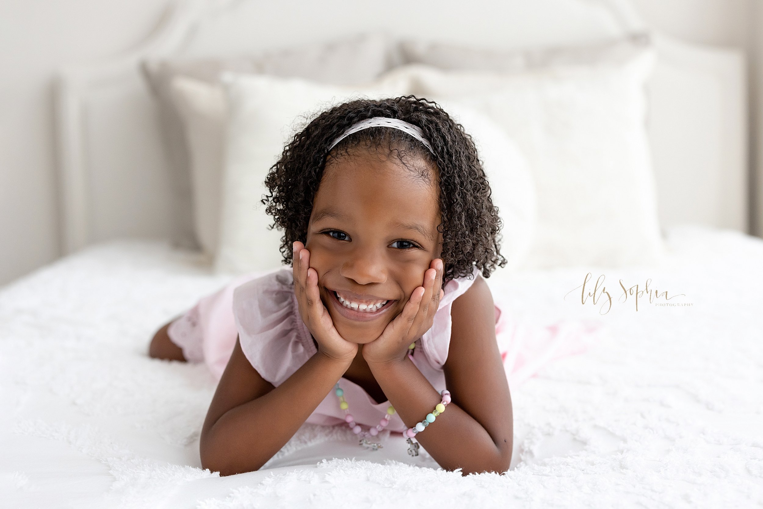  Family photo session with a three year old African-American girl as she lies on a bed with her hands on her cheeks taken in a natural light studio near Old Fourth Ward in Atlanta. 