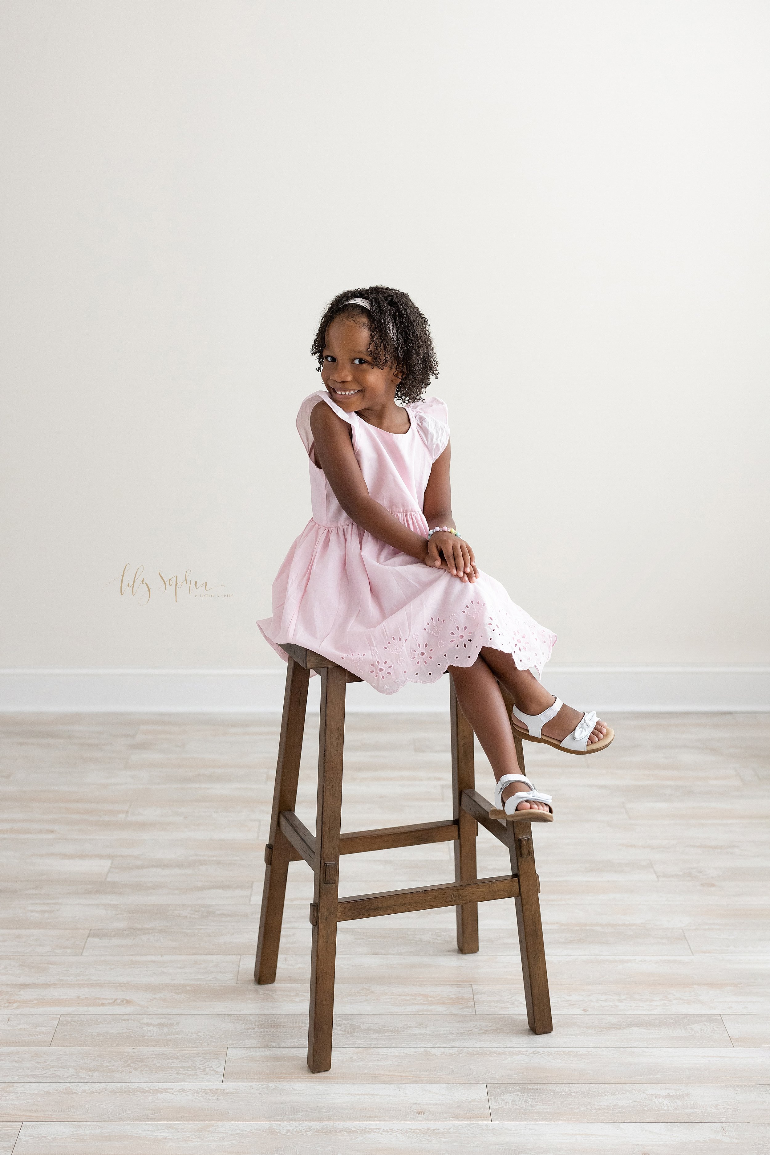  Family photo shoot of a young African-American girl as she sits with her legs crossed on a stool in front of a window streaming natural light places her hands on her knees and grins as she looks over her right shoulder taken near Oakhurst in Atlanta
