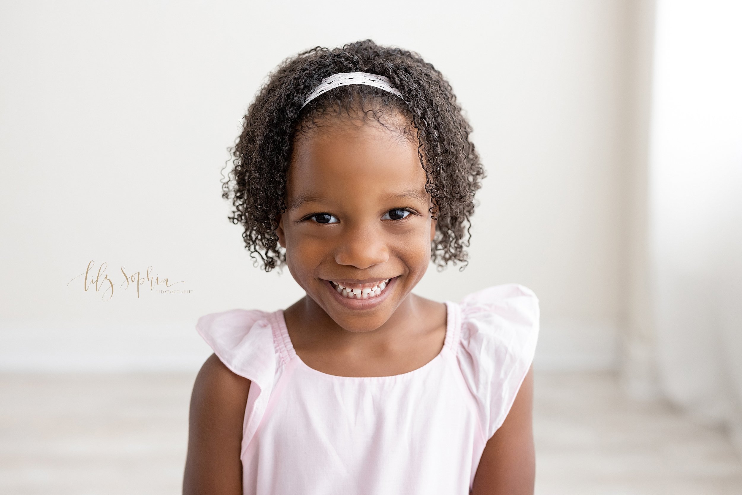  Family photo of a three year old smiling African-American girl as she stands in front of a window streaming natural light in a photography studio in Ponce City Market in Atlanta, Georgia. 