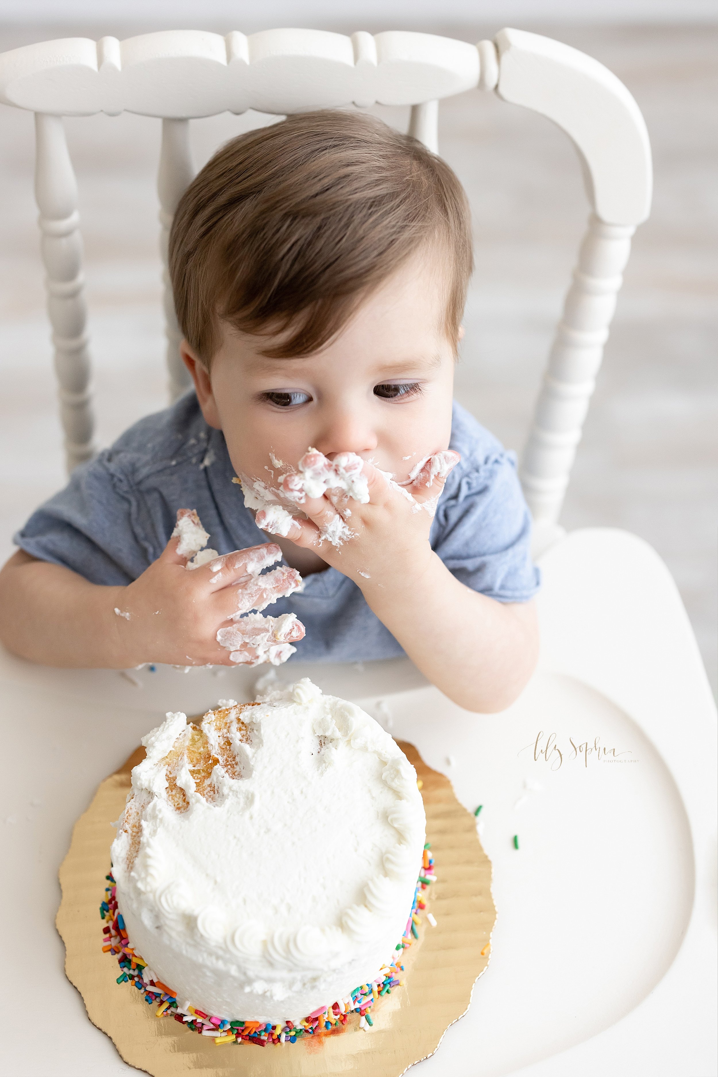  Smash cake first birthday portrait of a one year old boy licking his icing covered hands as he sits with his smash cake on the tray of the antique highchair in a studio near Decatur that uses natural light. 