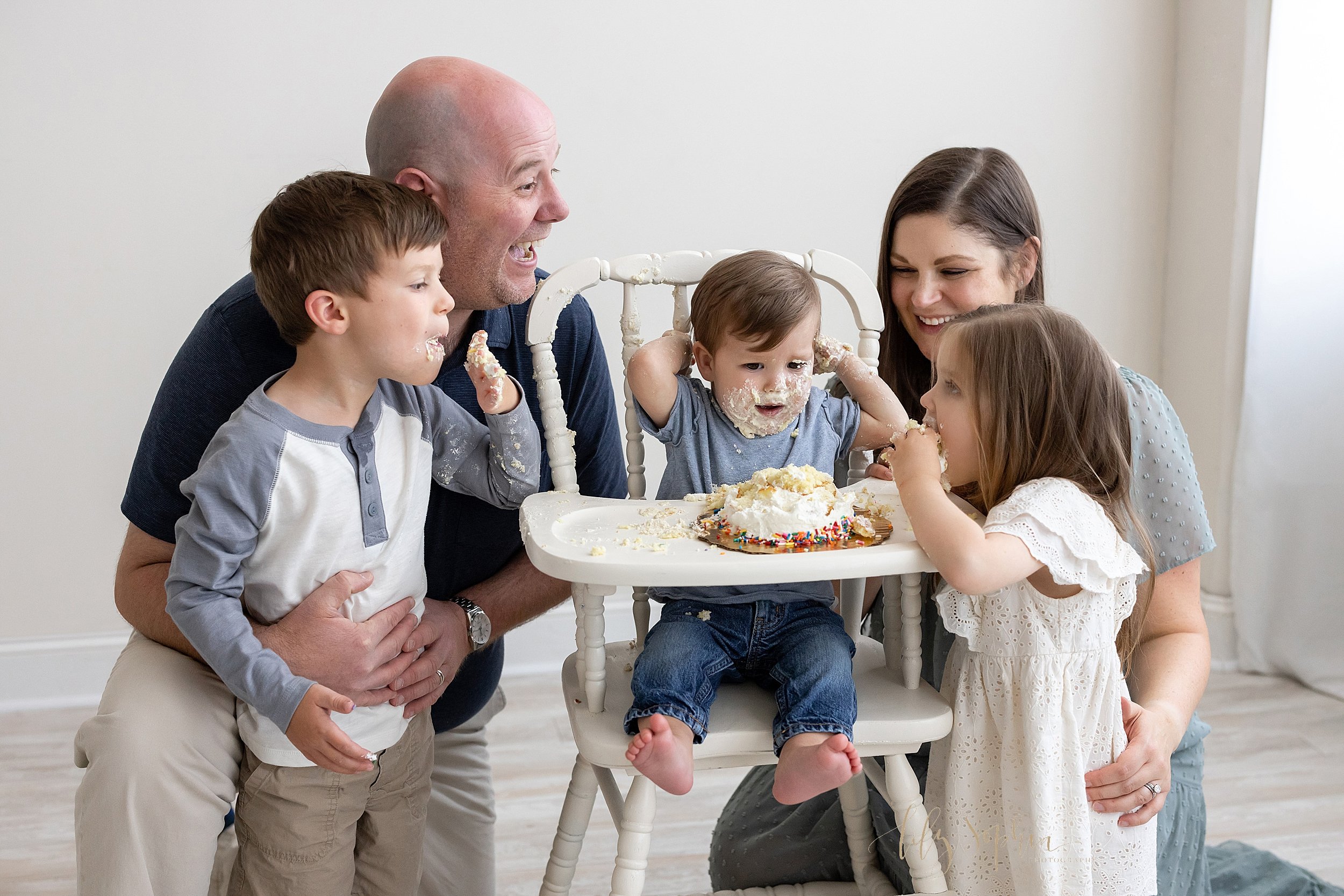  Fun family picture of a family of a one year old celebrating his first birthday with a smash cake as the birthday boy and his sibling devour it with mom and day watching taken in front of a window streaming natural light in a photography studio near