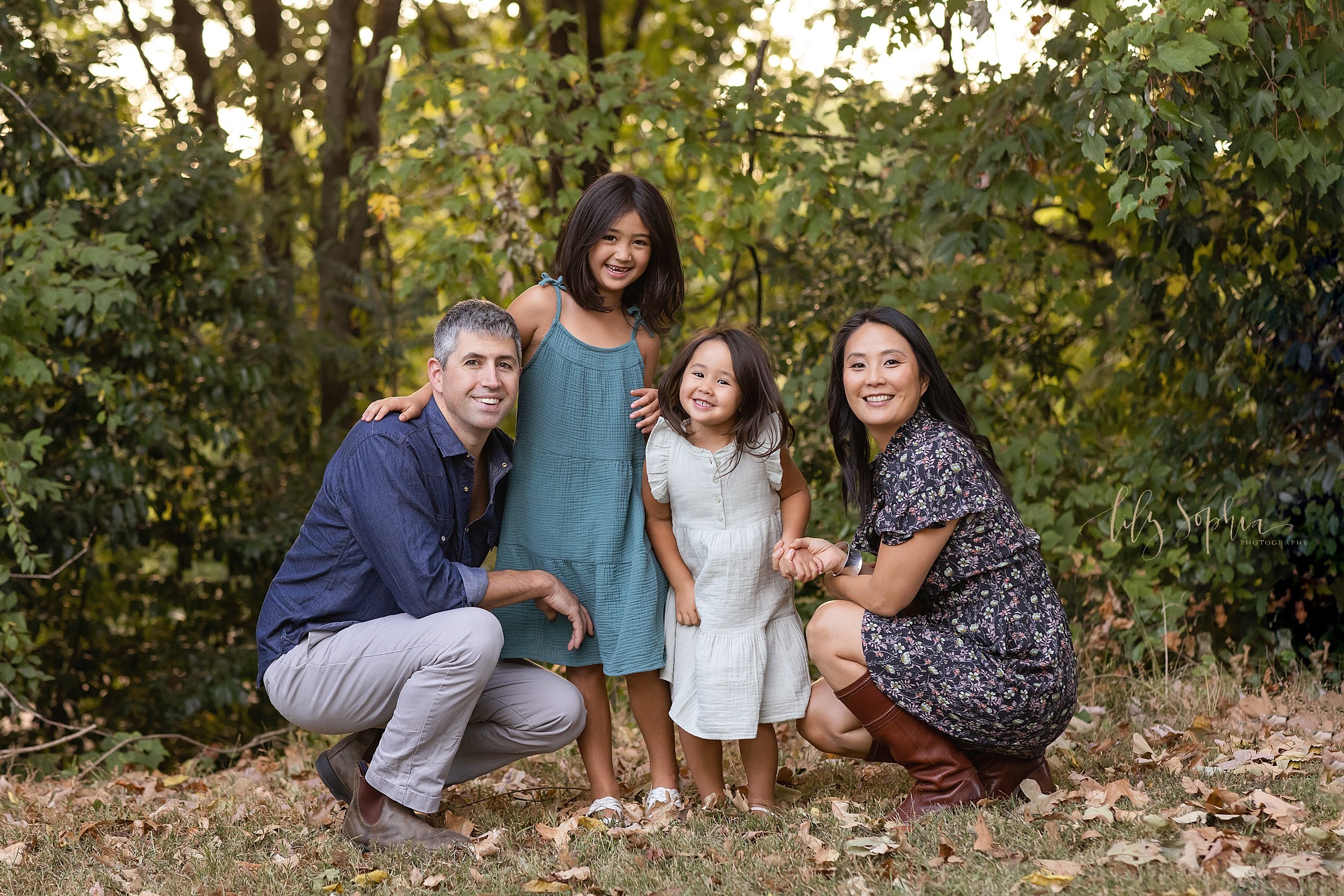  Family photo session of a mother and father squatting with their daughters between them in a park during autumn at sunset in Atlanta, Georgia. 