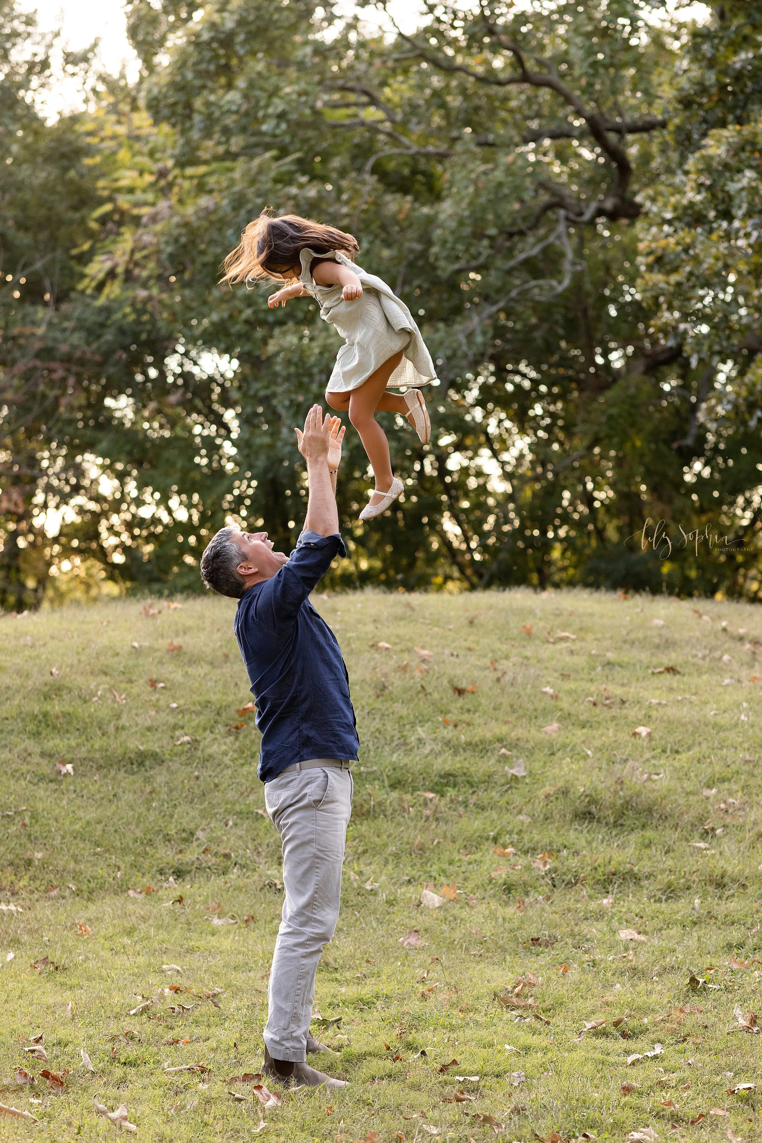  Fun family photo memory of a father throwing his young daughter in the air above his head as she flies at sunset in a park in Atlanta during autumn. 