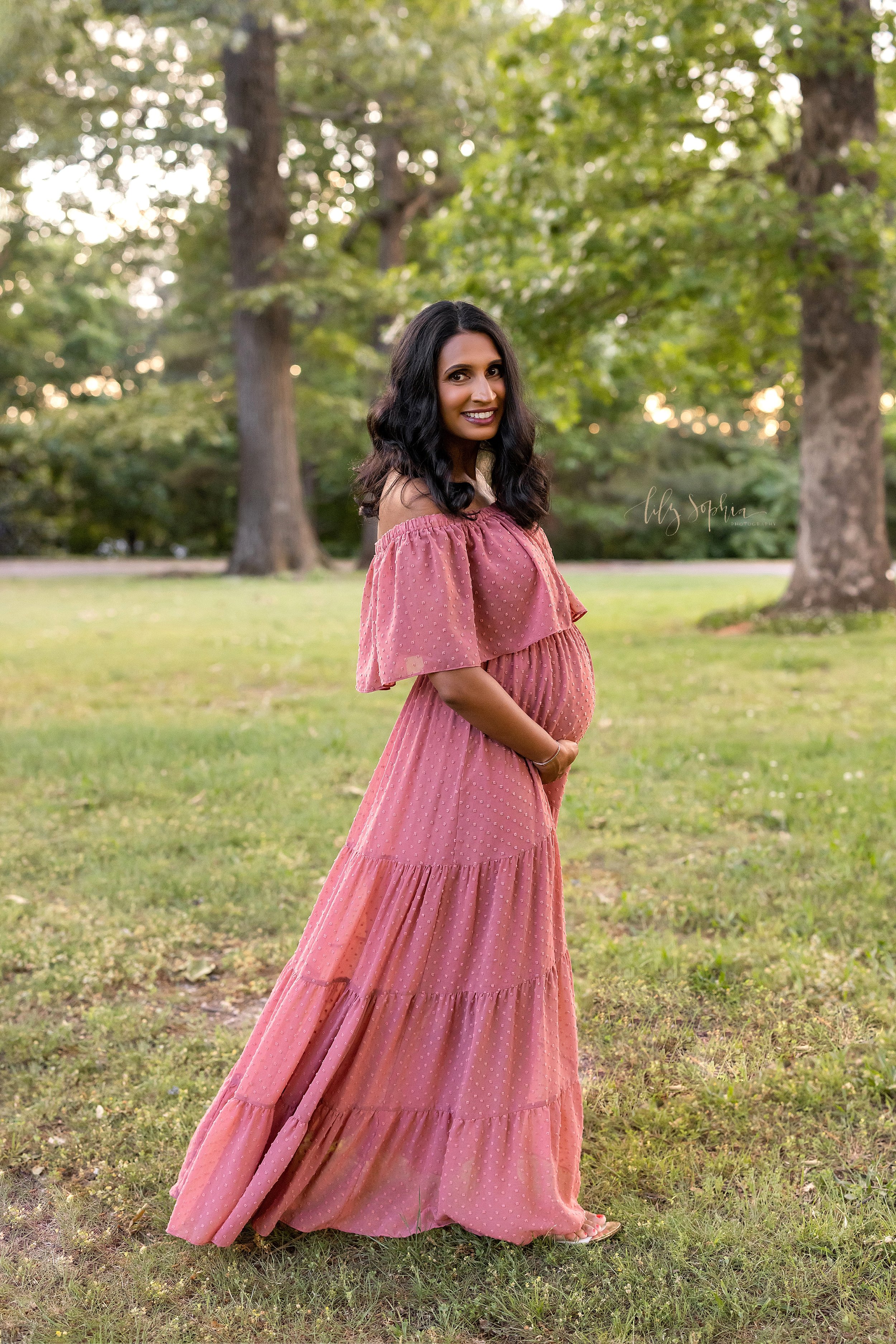  Maternity picture of an Indian mother wearing a flocked wide-bodiced off the shoulder ruffled full-length gown as she stands facing right holding her belly at sunset in an Atlanta park. 