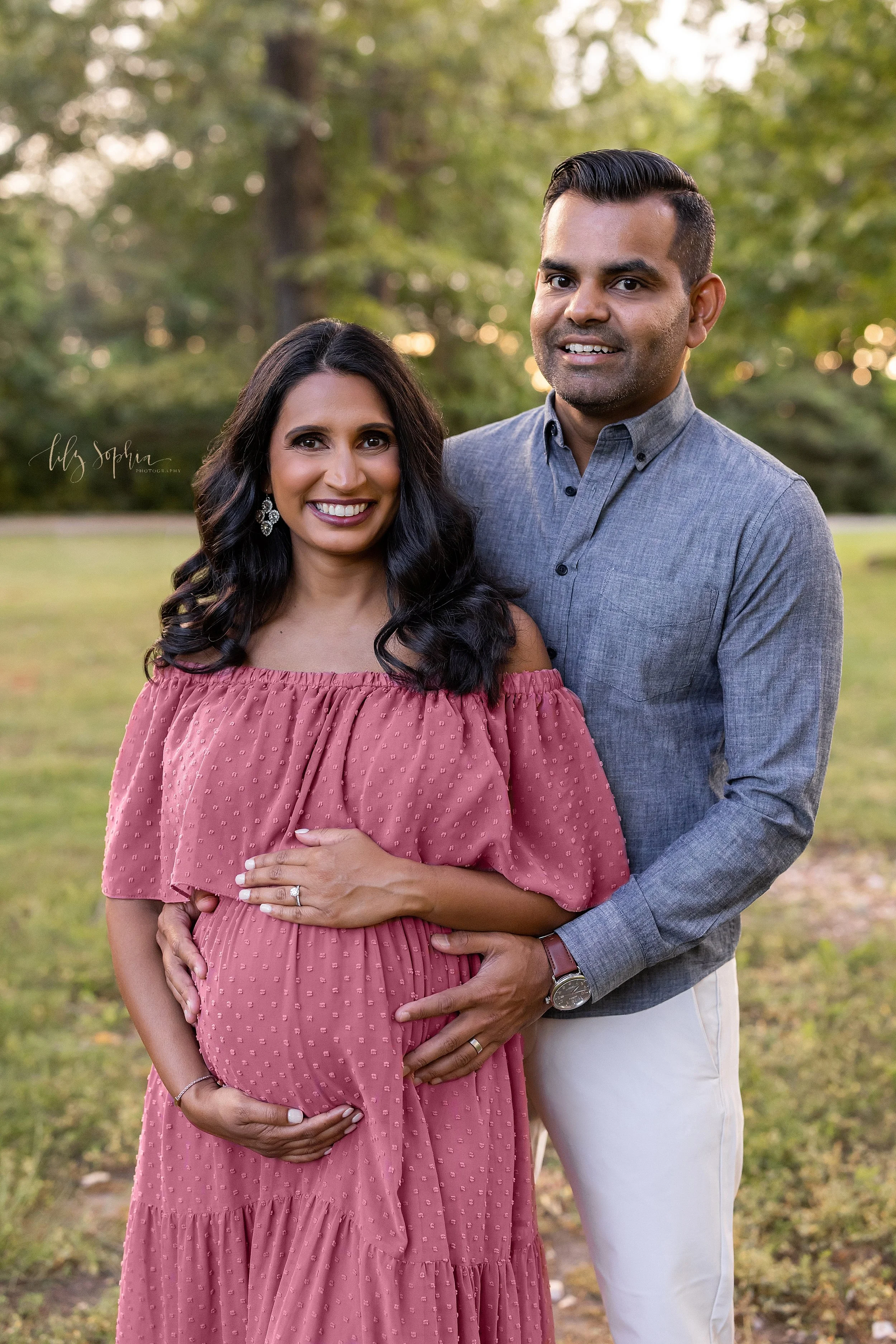  Maternity portrait of an Indian expectant couple as they stand at sunset in an Atlanta park with the wife framing her belly and the husband behind her placing his hand on her hip. 