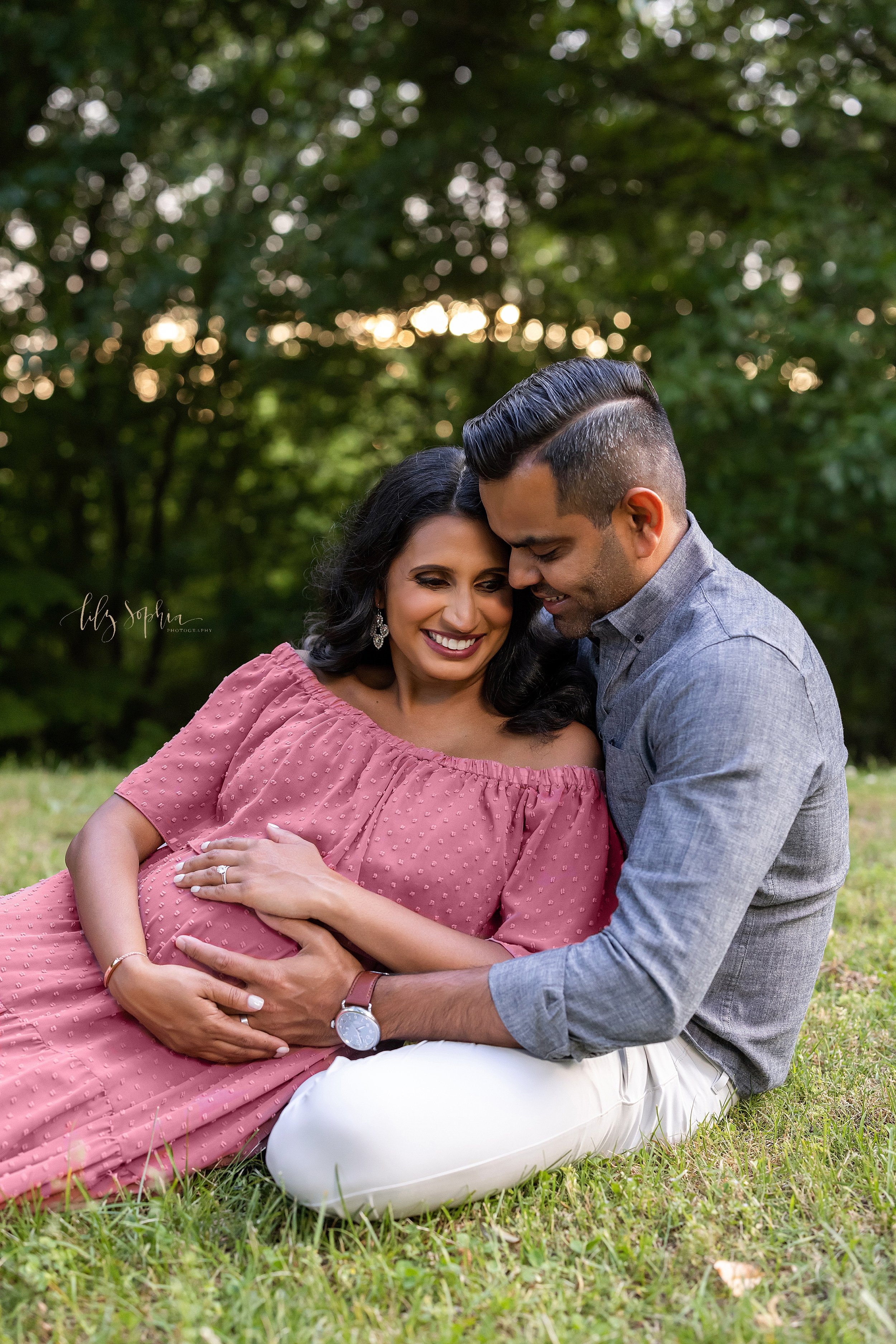  Maternity photo session in an Atlanta park with a pregnant mother lounging against her husband as they sit on the grass and feel the baby kicking in the womb at sunset. 