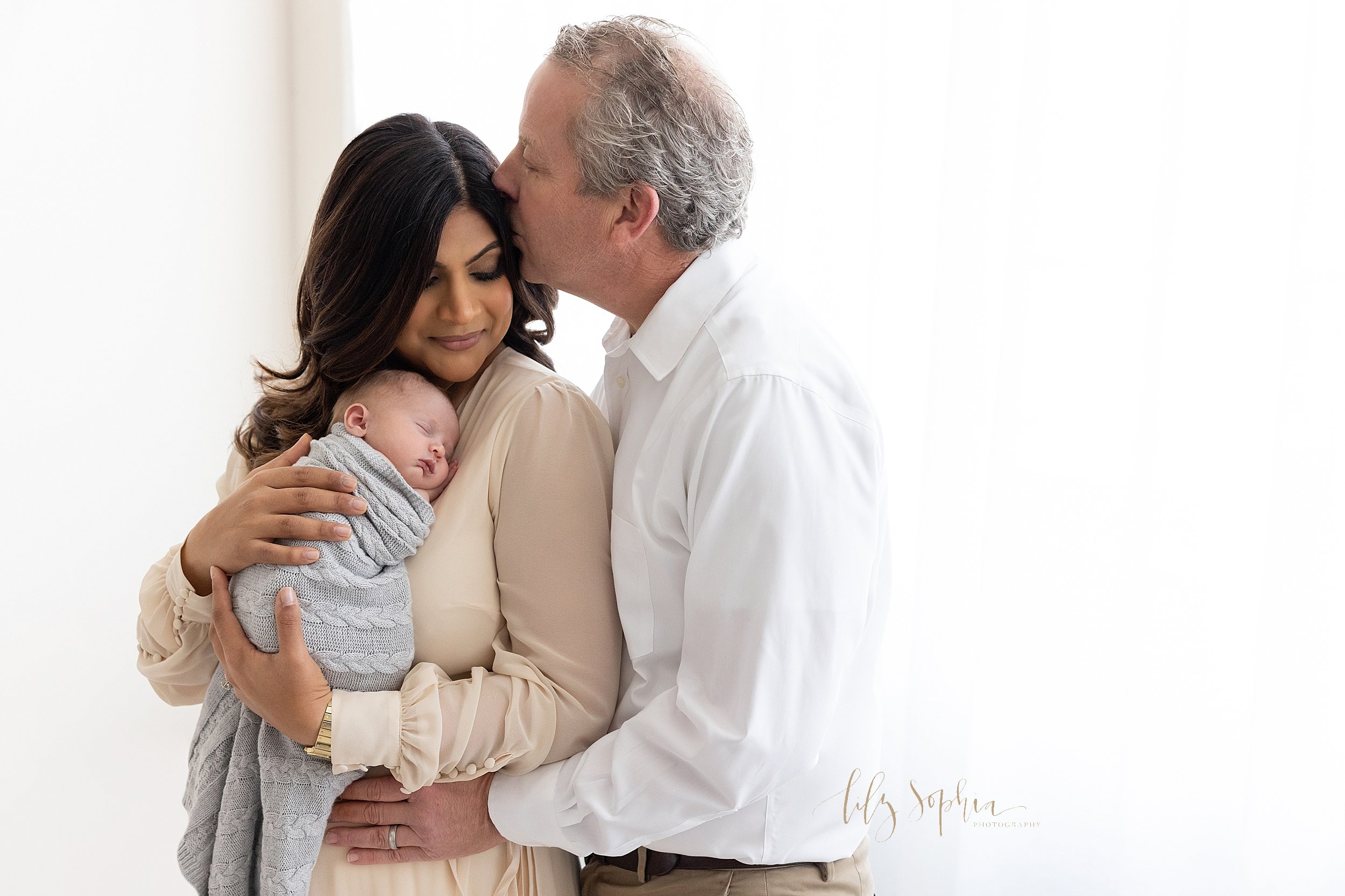  Family newborn portrait of a mother holding her newborn baby boy against her chest as her husband stand to her left side and holds her waist while kissing her on her head as the couple stand in front of a window streaming natural light in a photogra
