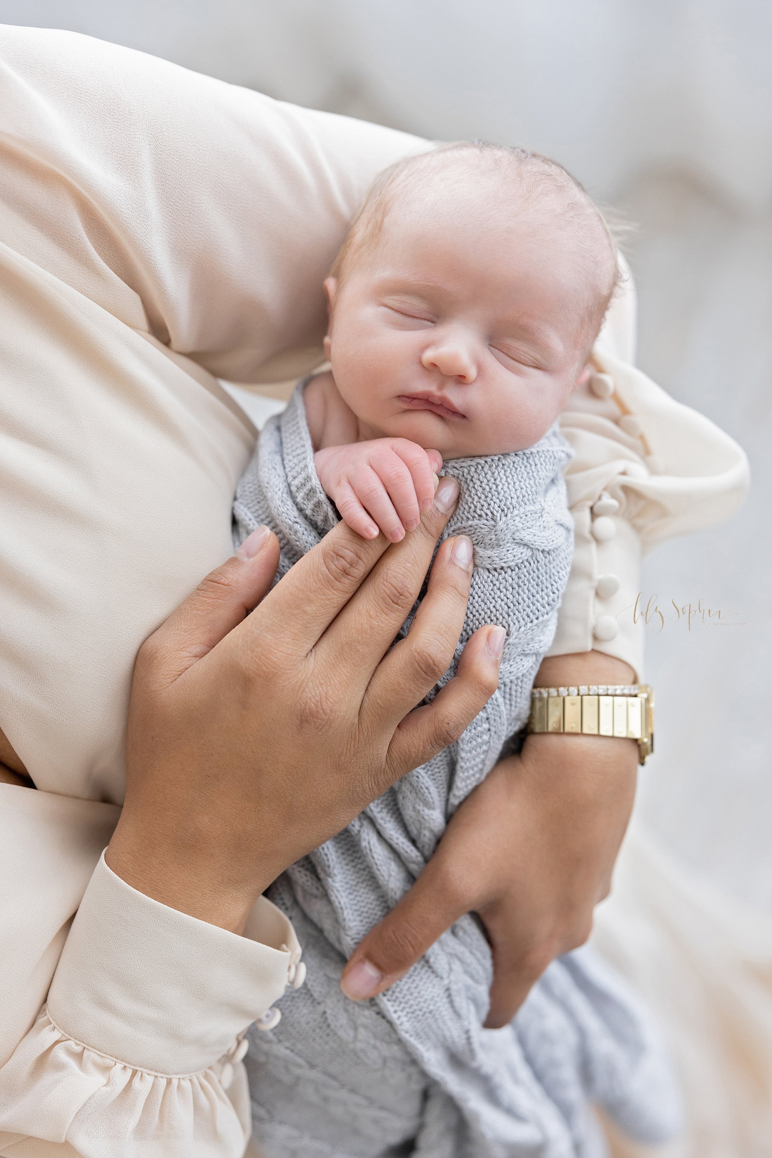  Newborn picture of a newborn baby boy as he holds on to his mother’s index finger while he peacefully sleeps taken in a studio that uses natural light near Buckhead in Atlanta, Georgia. 
