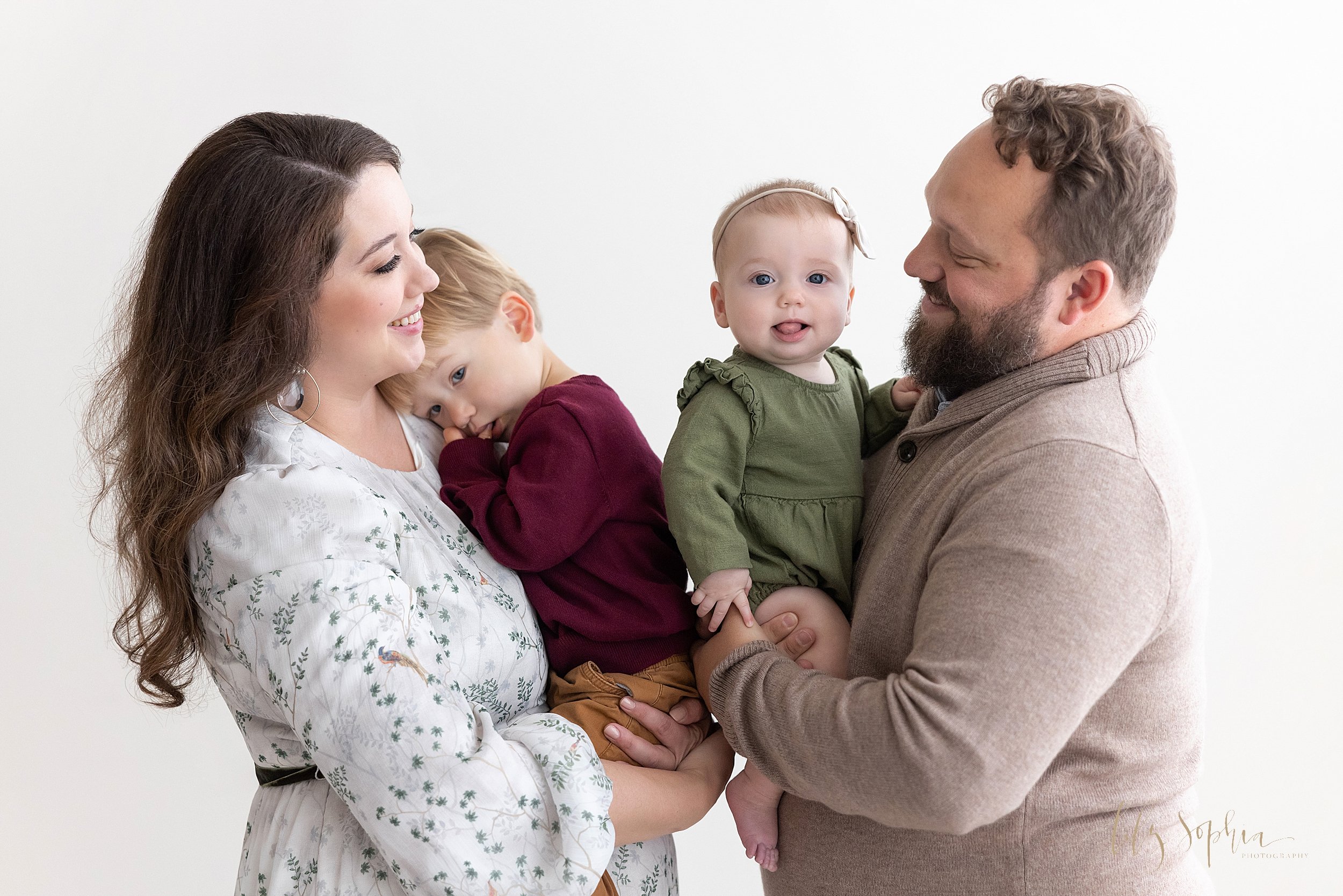  Family portrait of a mother holding their sleepy young son in her arms as he rests his head on her shoulder and dad holding their wide awake six month old daughter in his arms as the couple stands smiling at one another taken near Alpharetta in Atla