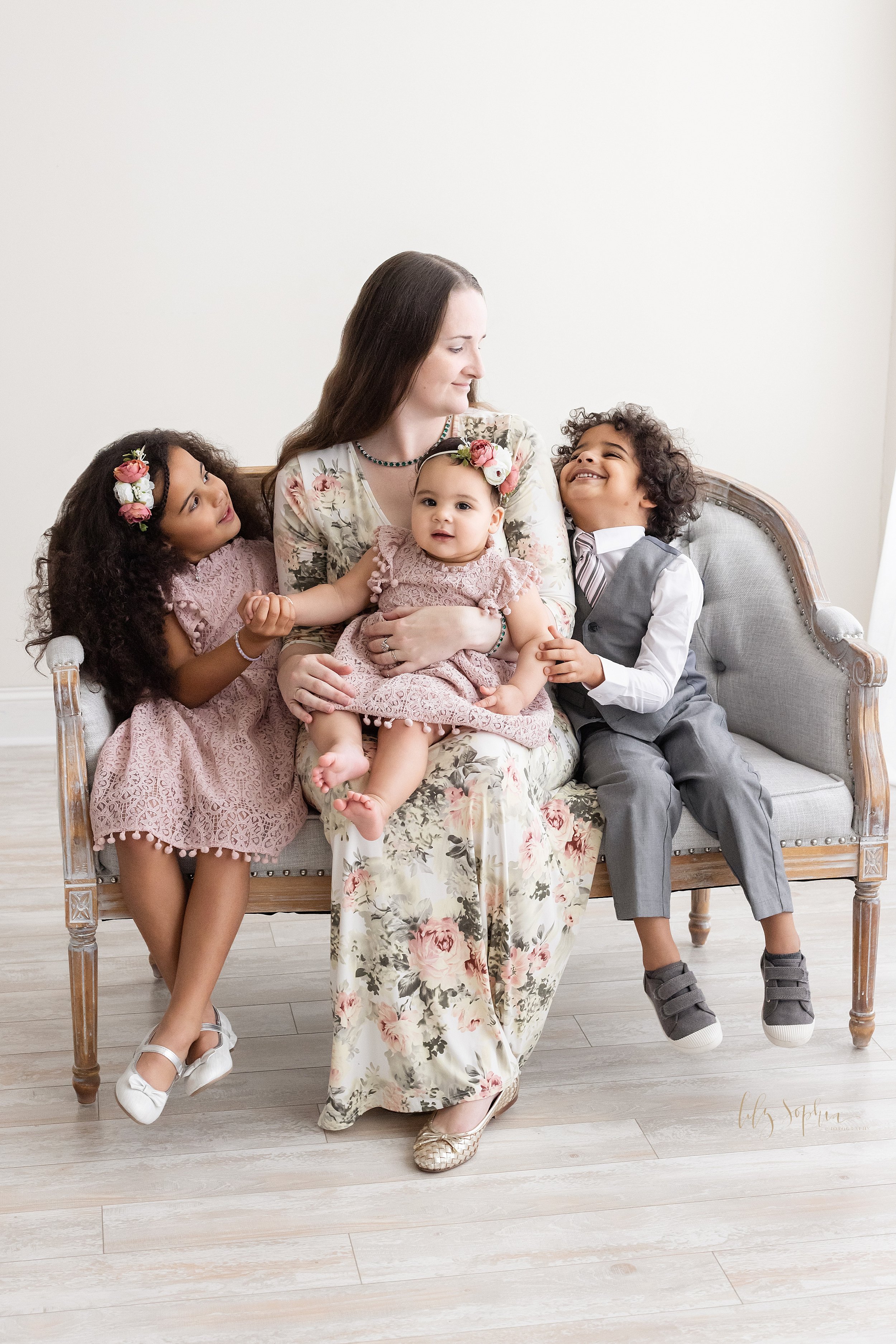  Family first birthday picture of a mother sitting in the middle of a sofa with her one year old daughter on her lap as her older daughter sits on her right side and holds her baby sister’s hand with her right hand and her son sits on her right side 