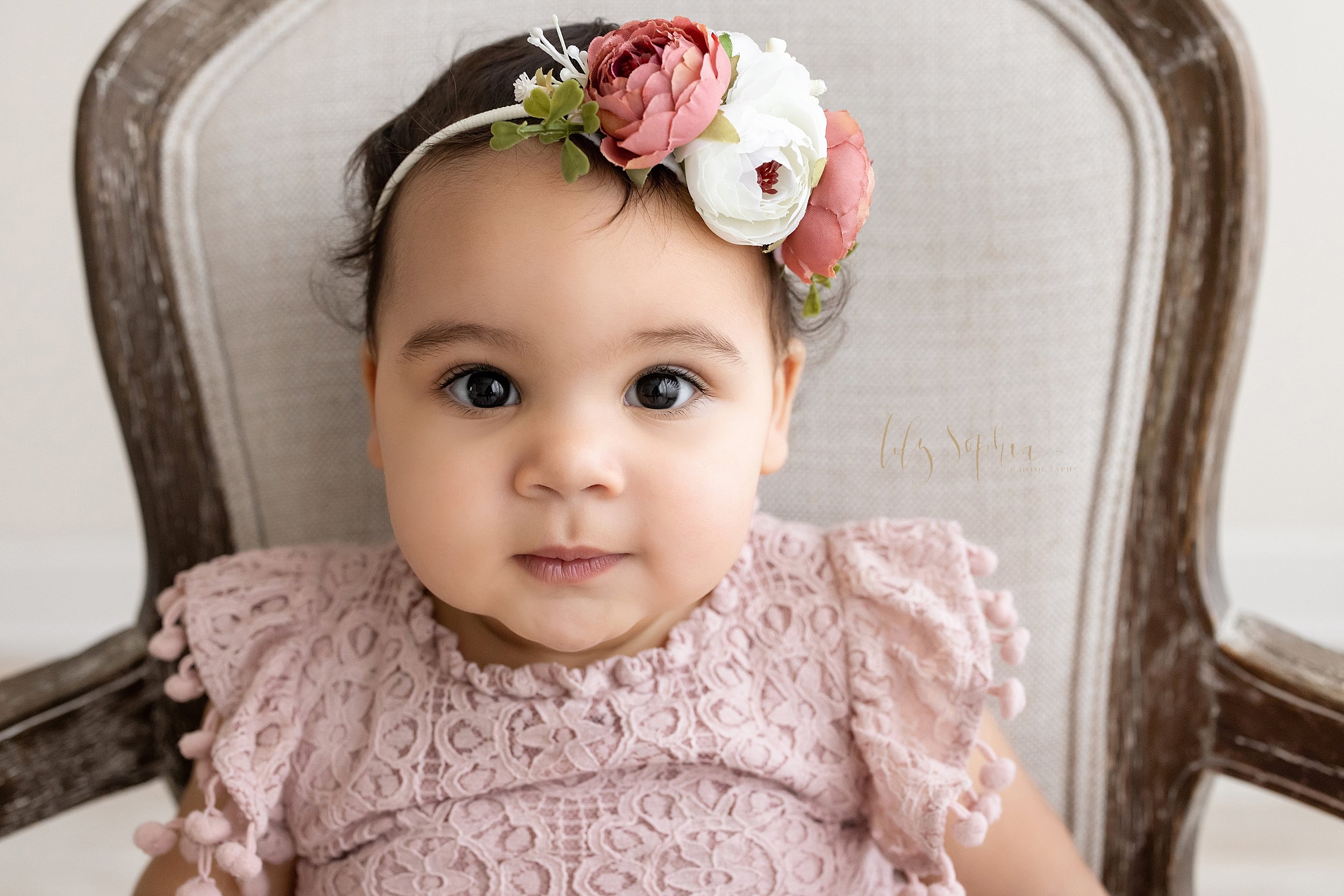  Close-up first birthday portrait of a one year old baby girl sitting in a chair wearing a headband with large flowers on it taken in a natural light studio near Kirkwood in Atlanta, Georgia. 