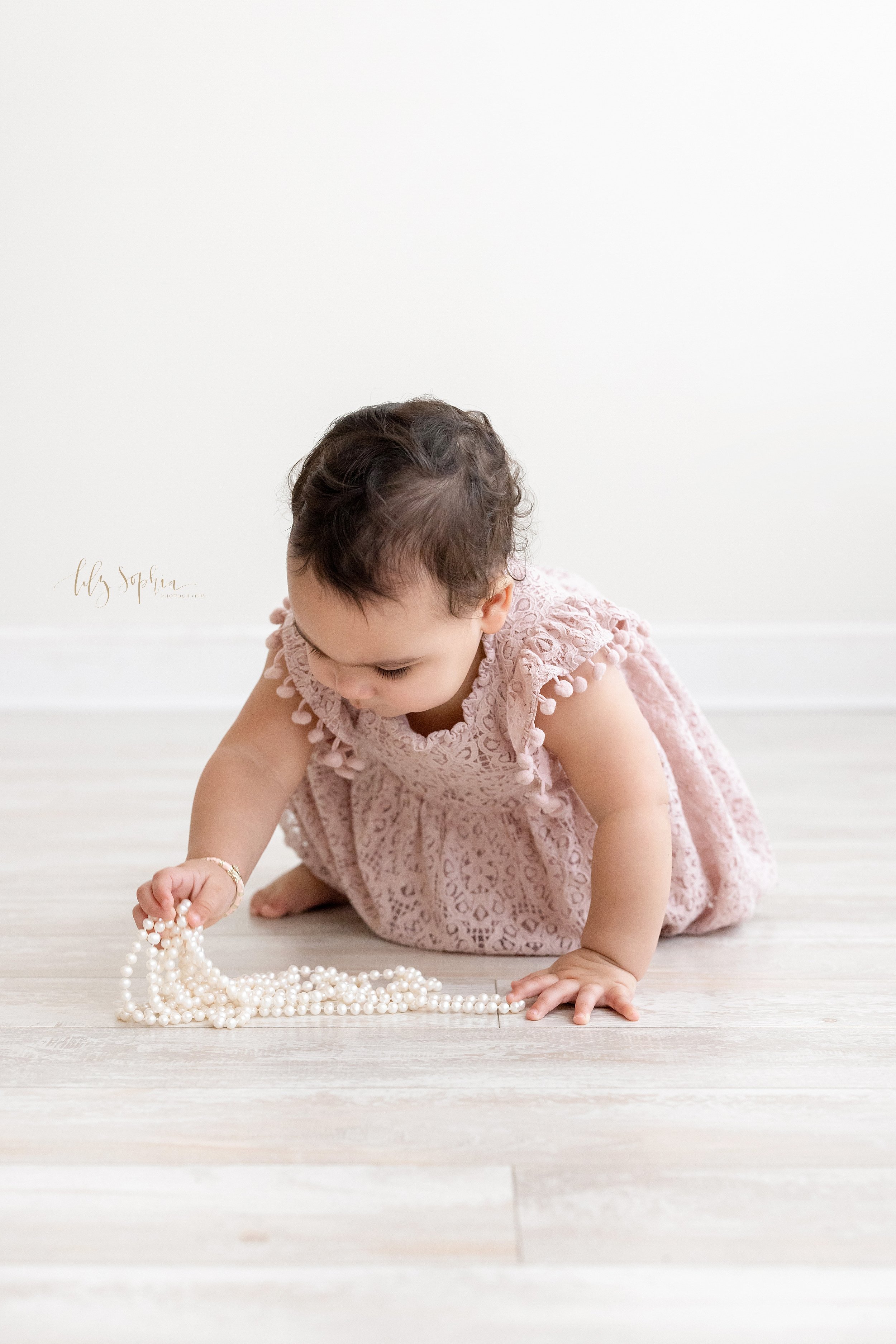  First birthday memory portrait of a one year old baby girl crawling on the floor of a photography studio to play with her mother’s string of pearls taken near Poncey Highlands in Atlanta that uses natural light. 
