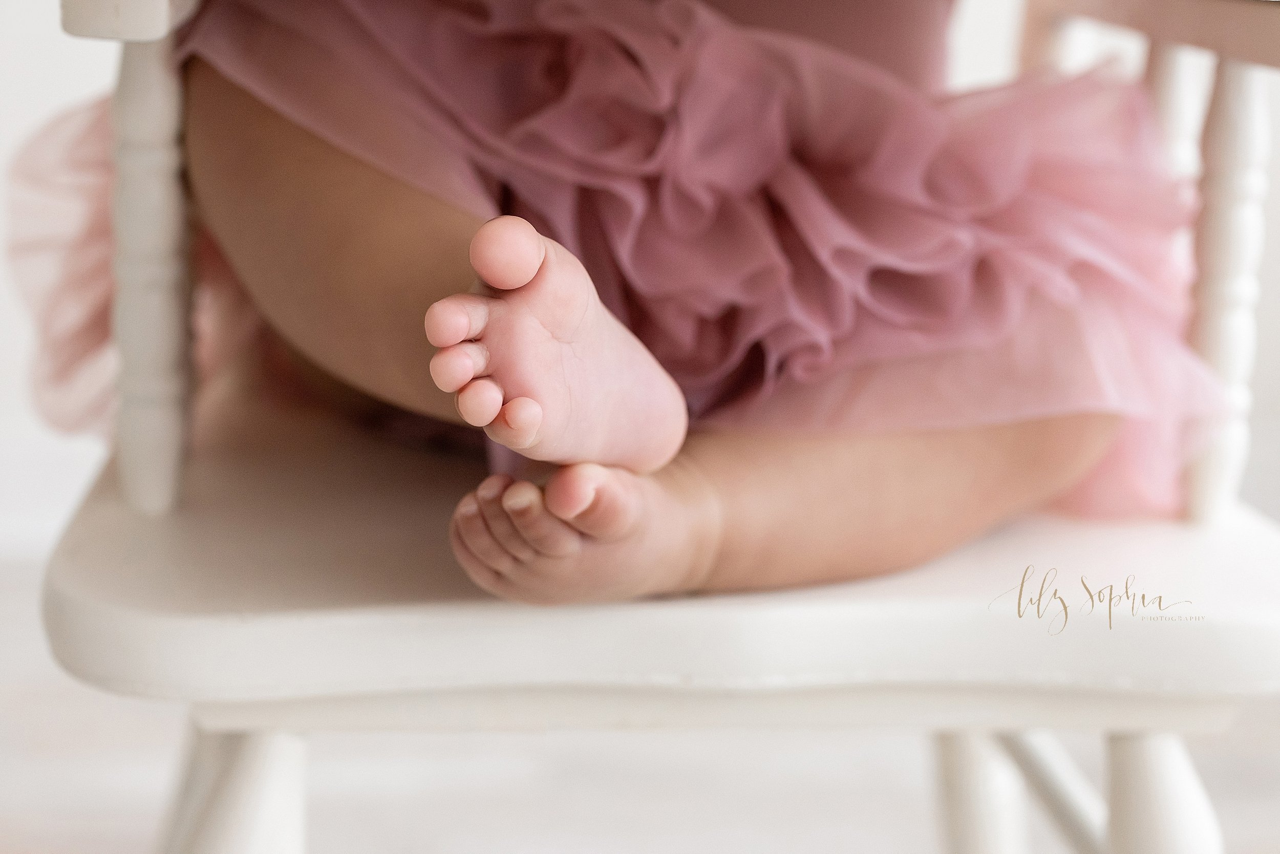  First birthday memory photo of a one year old girl’s tiny feet and toes as she sits in a highchair waiting for her smash cake taken in a studio near Ansley Park in Atlanta that uses natural light. 