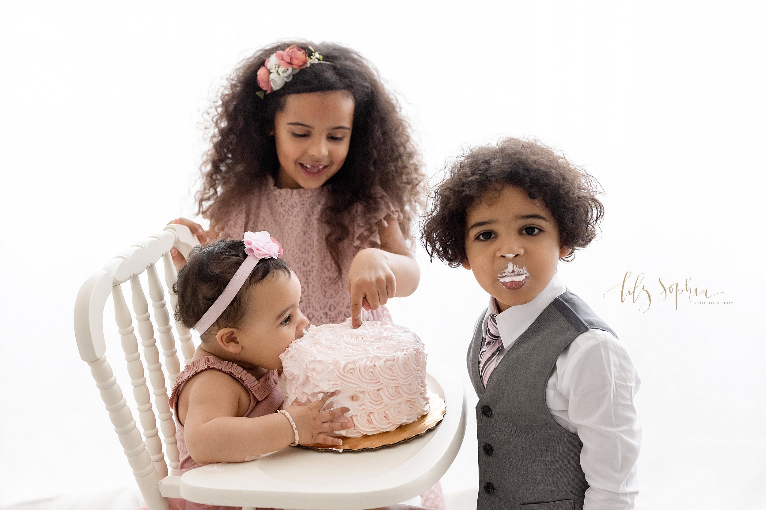  Smash cake first birthday family photo of a one year old baby girl sitting in an antique highchair holding her cake and taking a bite while her older sister stands next to her and put her finger into the frosting and her brother faces her while look