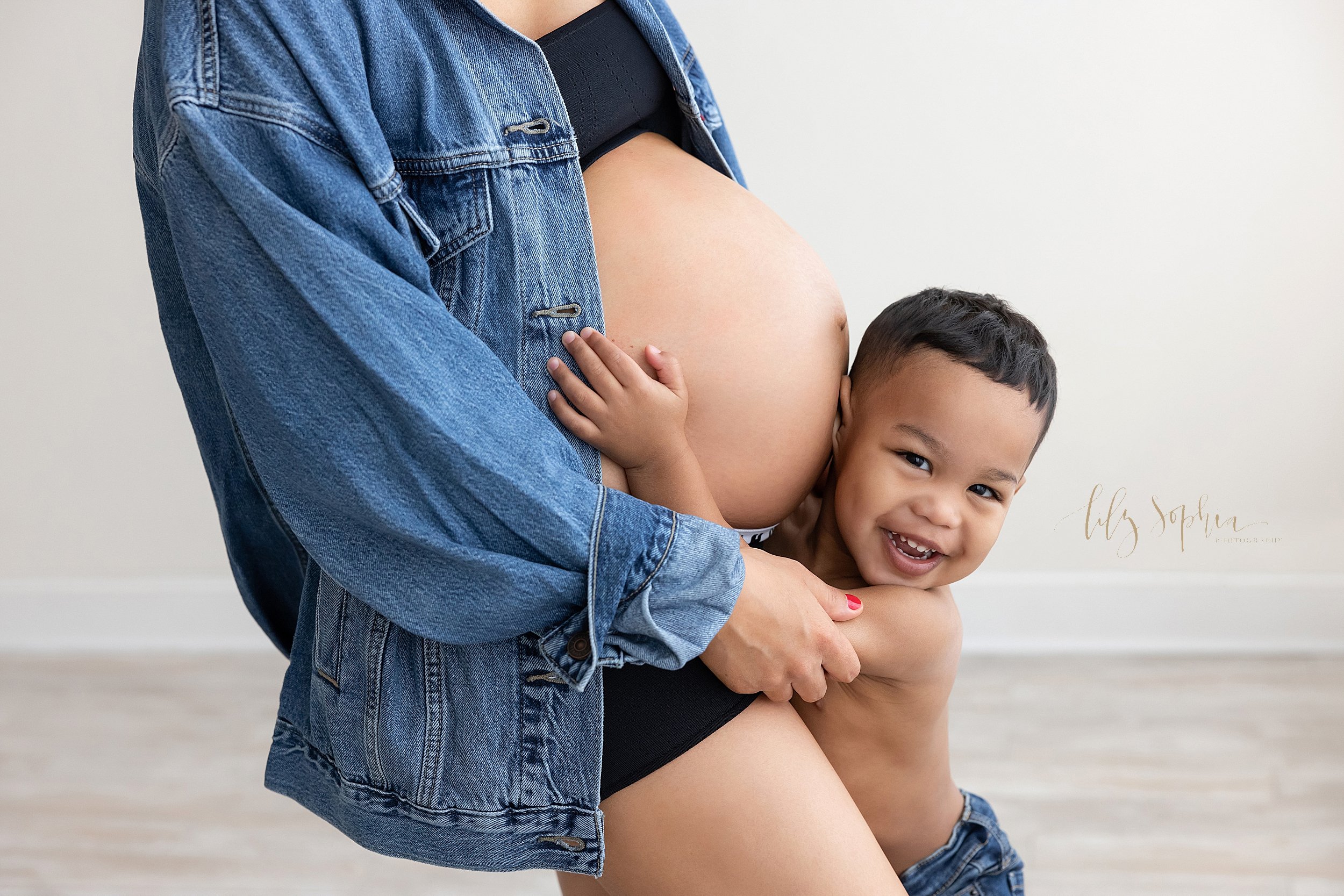  Maternity picture of a pregnant mother wearing a denim shirt, a sports bra, and stretchy shorts that allow her belly to be bare as her young son holds her belly and listens to his sibling in utero taken in a studio using natural light near Roswell i