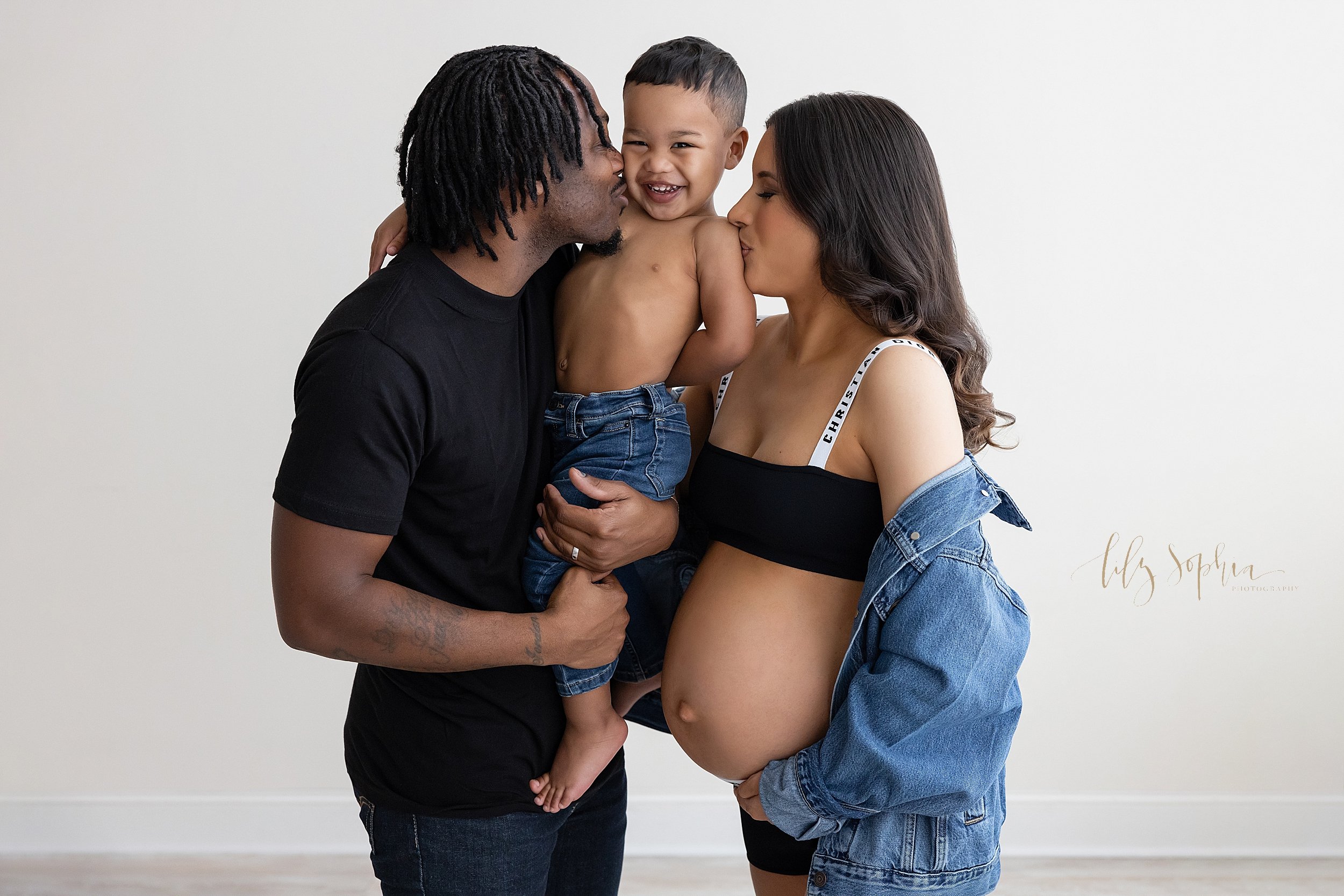  Family maternity photo of a father holding his young son in his arms as mom stands facing her husband with her belly bared and both parents kiss their son taken near Morningside in Atlanta in a natural light studio. 