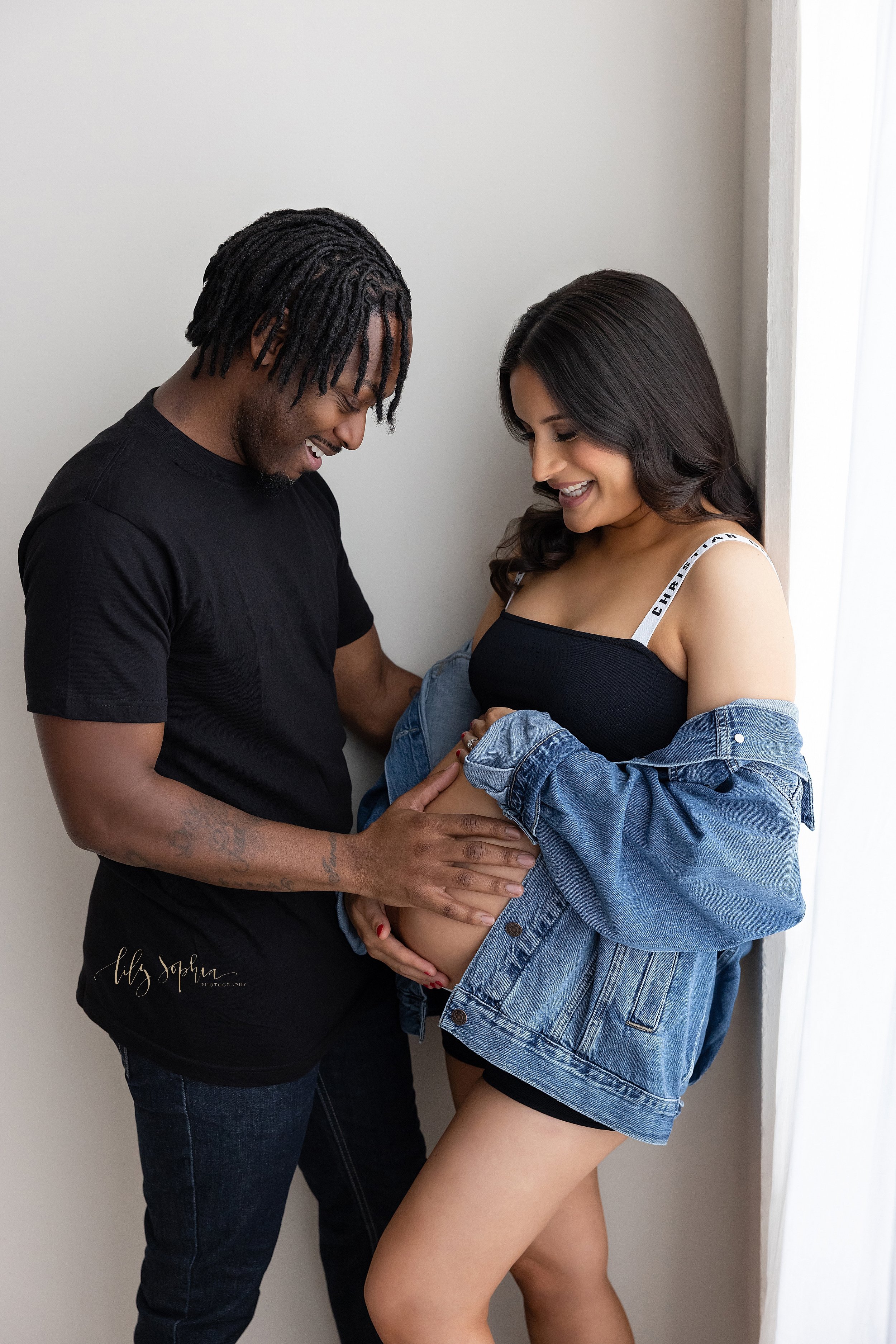  Maternity photo shoot of a pregnant mother standing next to a window streaming natural light as her husband faces her and places his hand on her bare belly to feel his child in utero moving taken near Virginia Highlands in Atlanta, Georgia. 