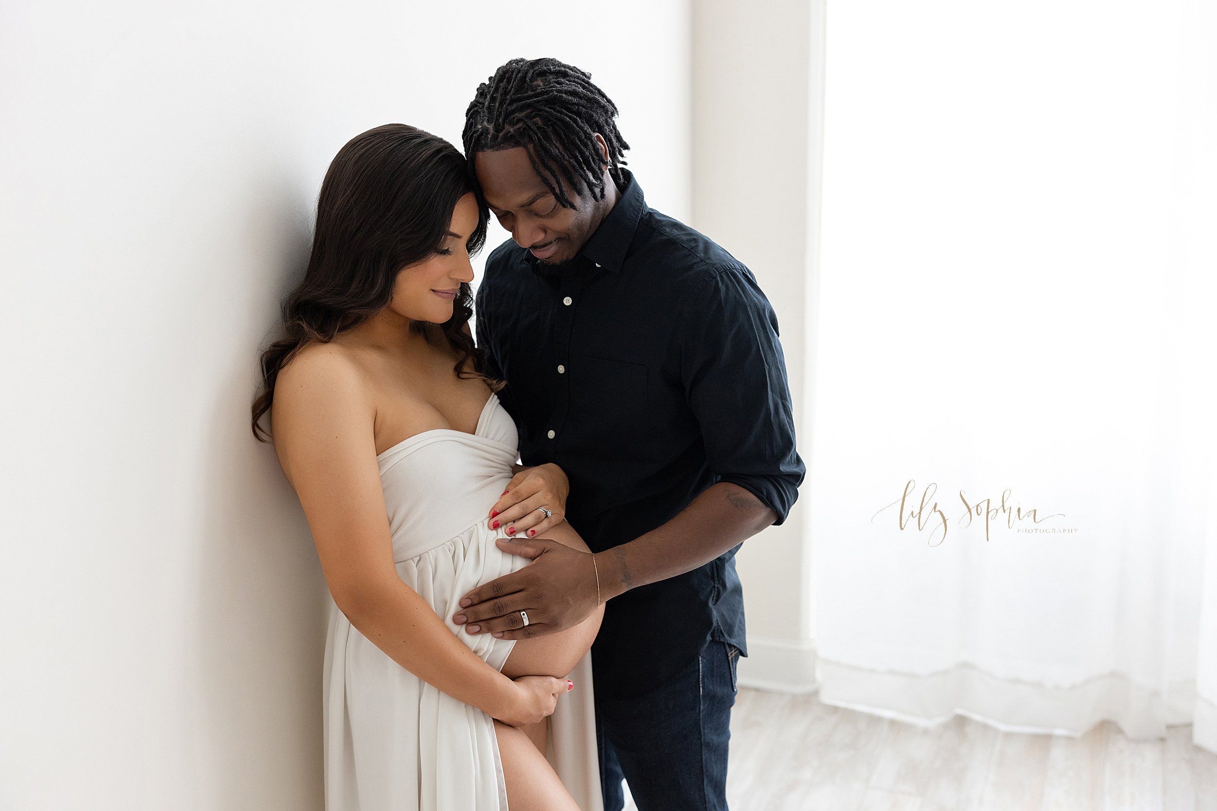  Maternity photo session with a pregnant mother wearing a strapless split-front gown  allowing her belly to be bared and framing it with her hands as she stands against a wall with her husband on her right side with his hand on their child in utero a