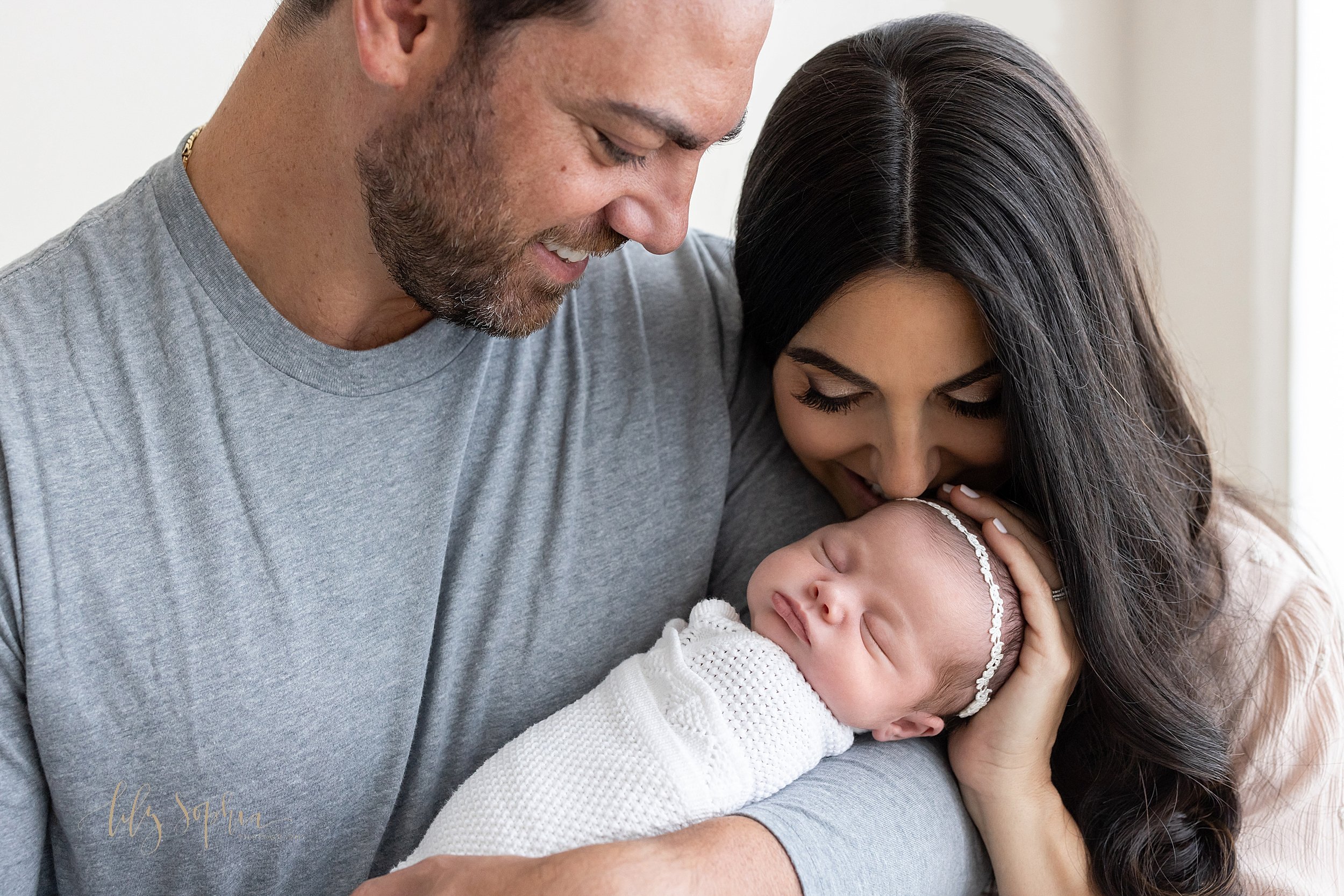  Newborn photo session of a father cradling his infant daughter in his arms while admiring her and his wife standing to his right side with her left hand on her daughter’s head as she leans down to talk with her as the couple stand next to a window s