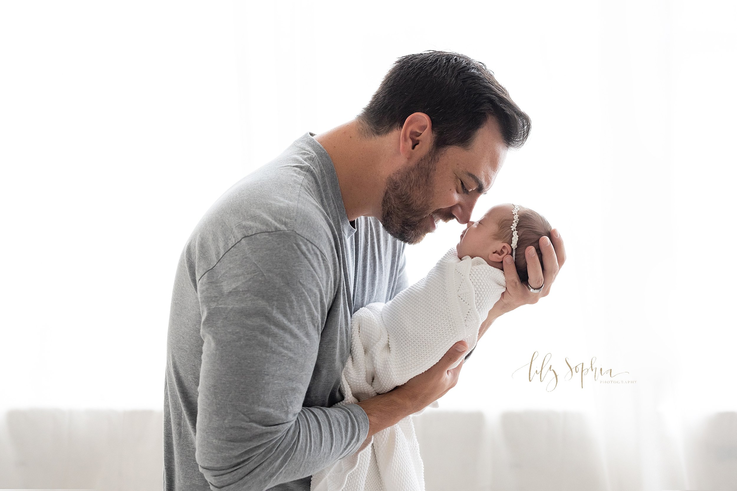  Newborn photo session in a natural light studio of a father standing and holding his newborn daughter in front of him in his hands as he touches noses with her and talks to her taken near Kirkwood in Atlanta. 