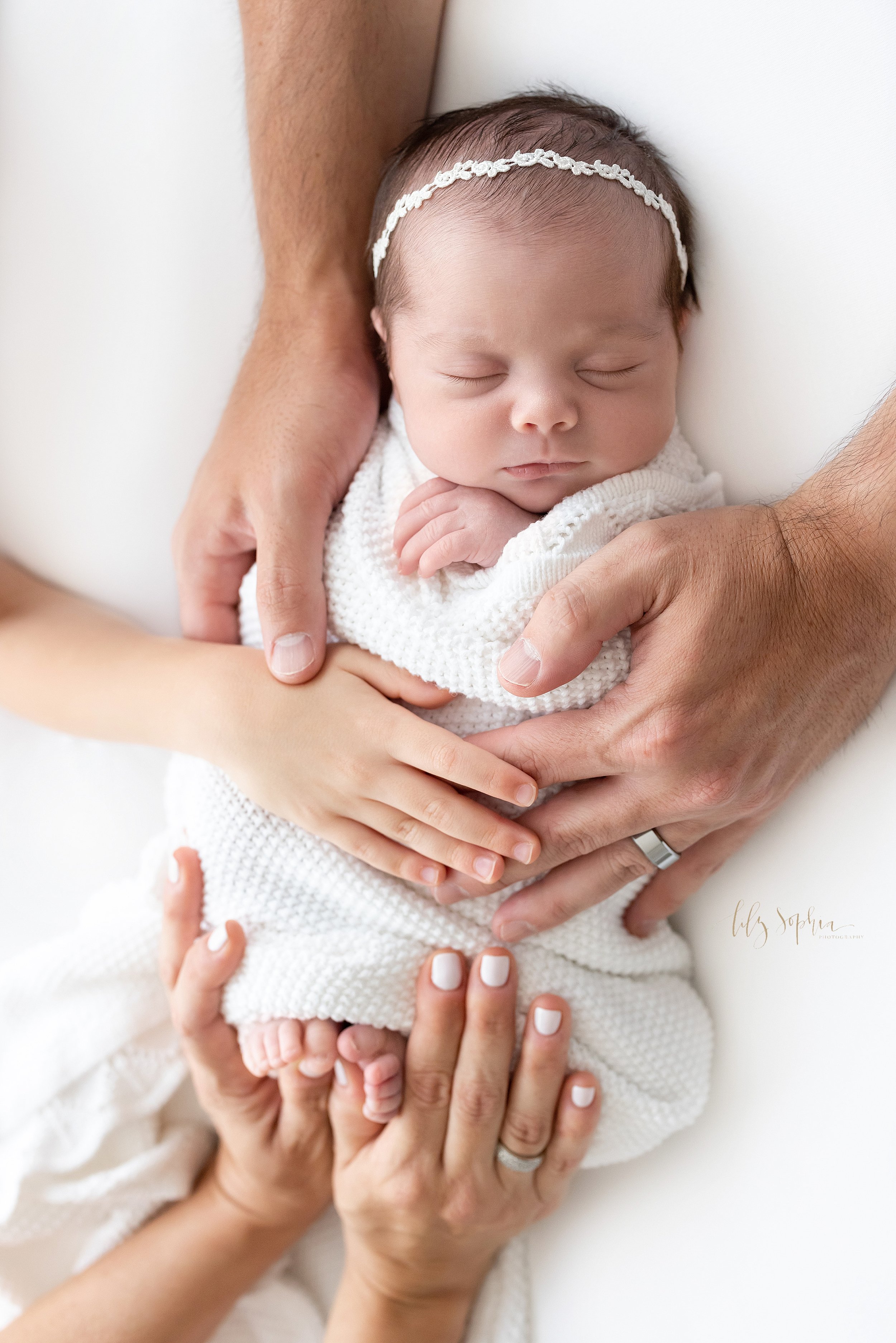  Newborn photo shoot in a studio near Cumming in Atlanta of a newborn baby girl lying on her back with her dad’s strong hands on her shoulders, her mother’s hands holding her feet and her brother’s hand on her body taken using natural light. 