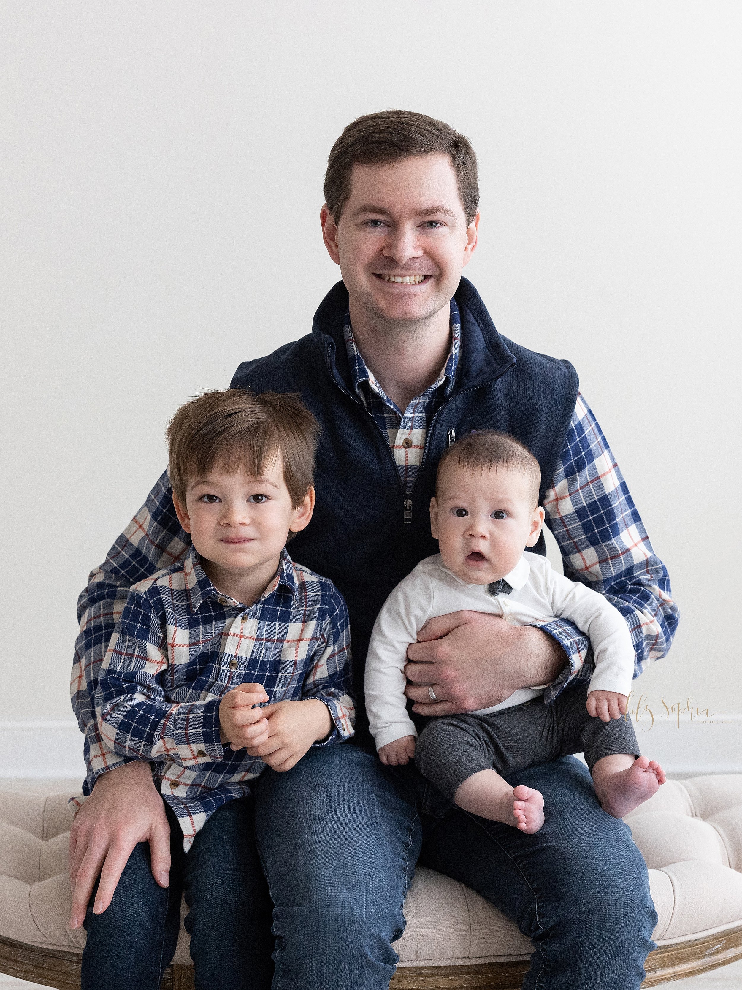  Family portrait of a father sitting on a tufted ottoman with his baby boy on his left knee as the baby turns his head to the left to look at his older brother who is sitting next to his father on his right side taken using natural light near Morning