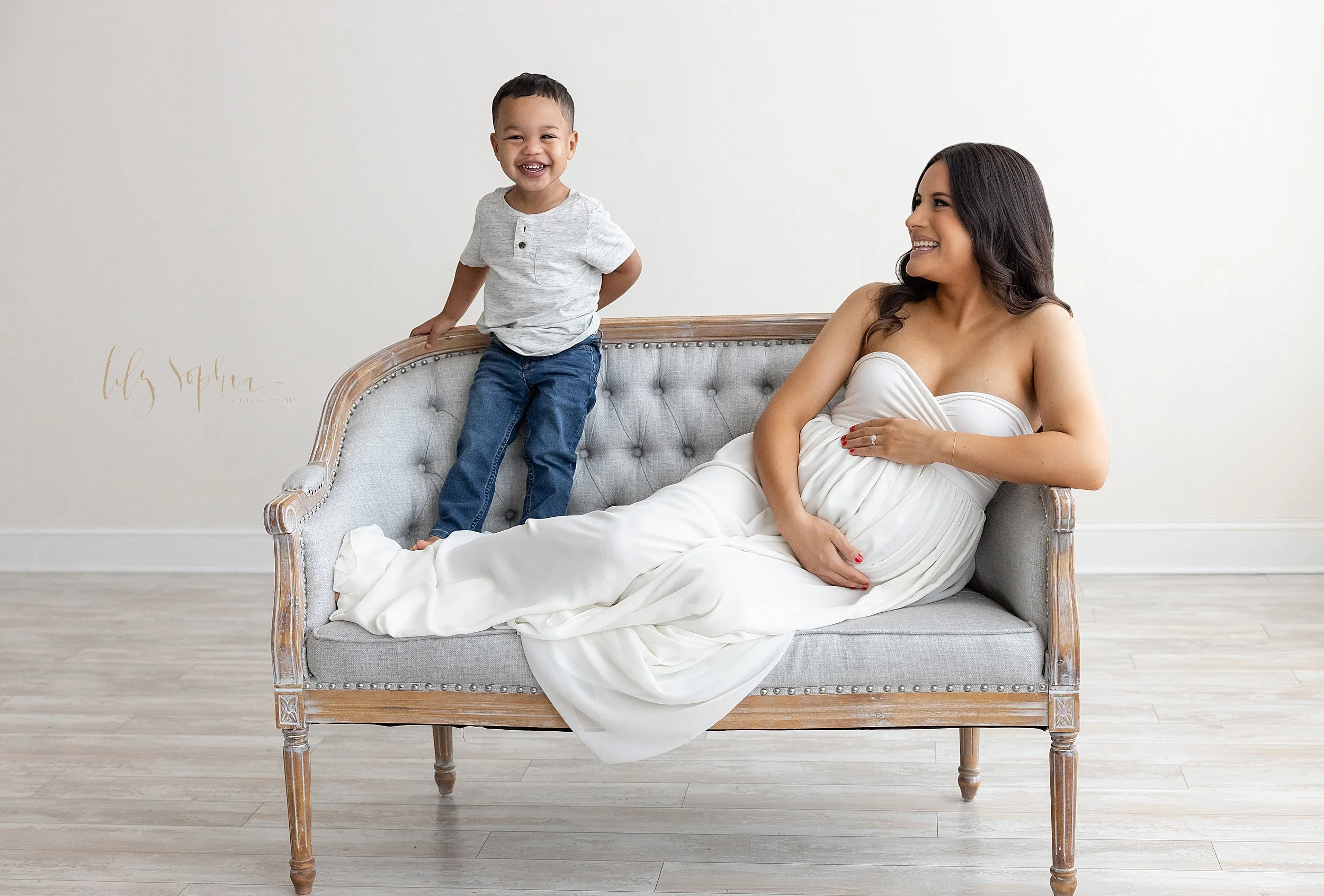  Maternity picture of a pregnant mother lounging on a tufted settee while framing her belly with her hands as her young son stands grinning and holding on to the settee back for balance taken in Ponce City Market in Atlanta in a natural light studio.