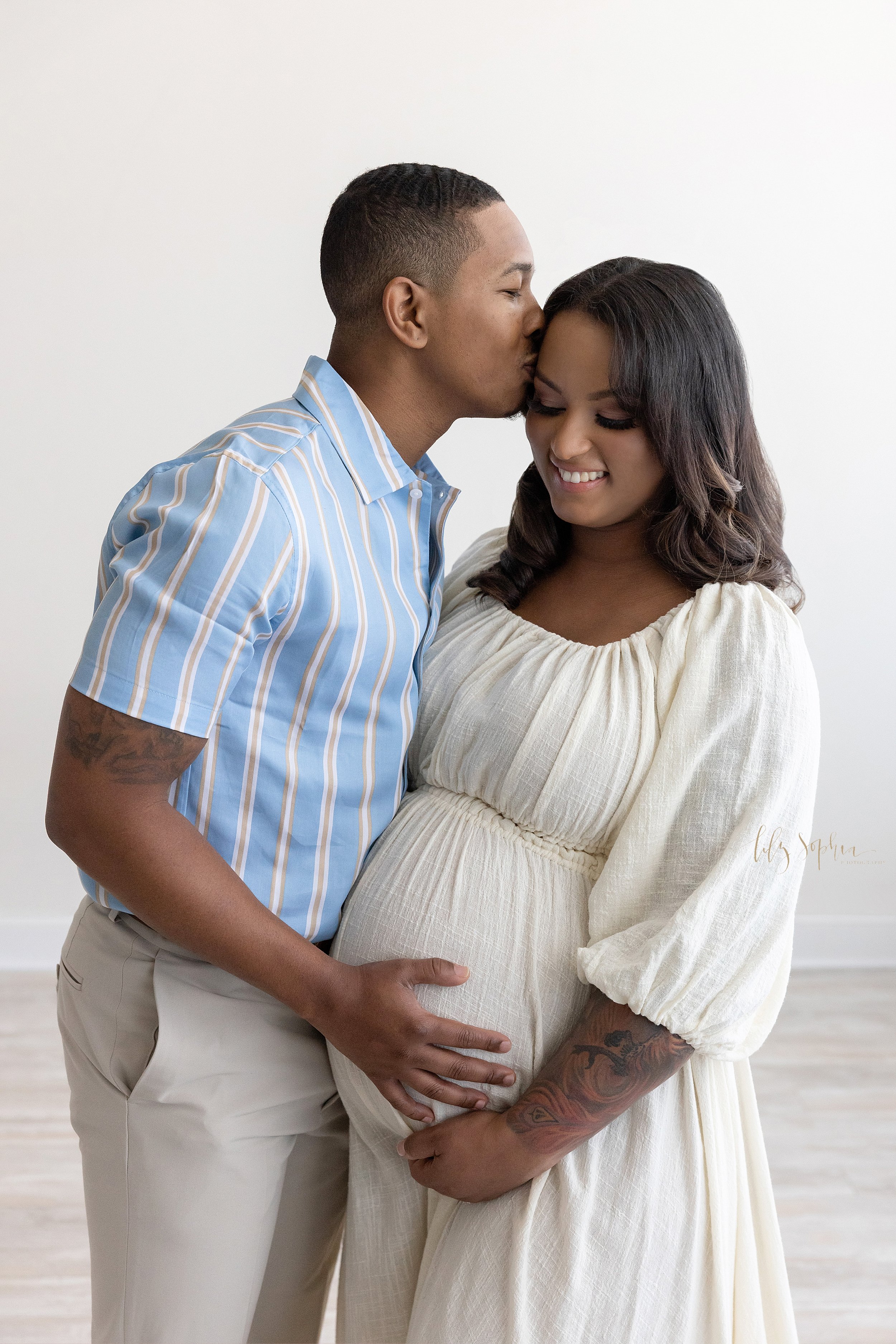  Maternity photo session with an African-American pregnant mother holding the base of her belly as her husband stands facing her with his right hand on their child in utero while kissing the side of his wife’s head taken near Old Fourth Ward in Atlan