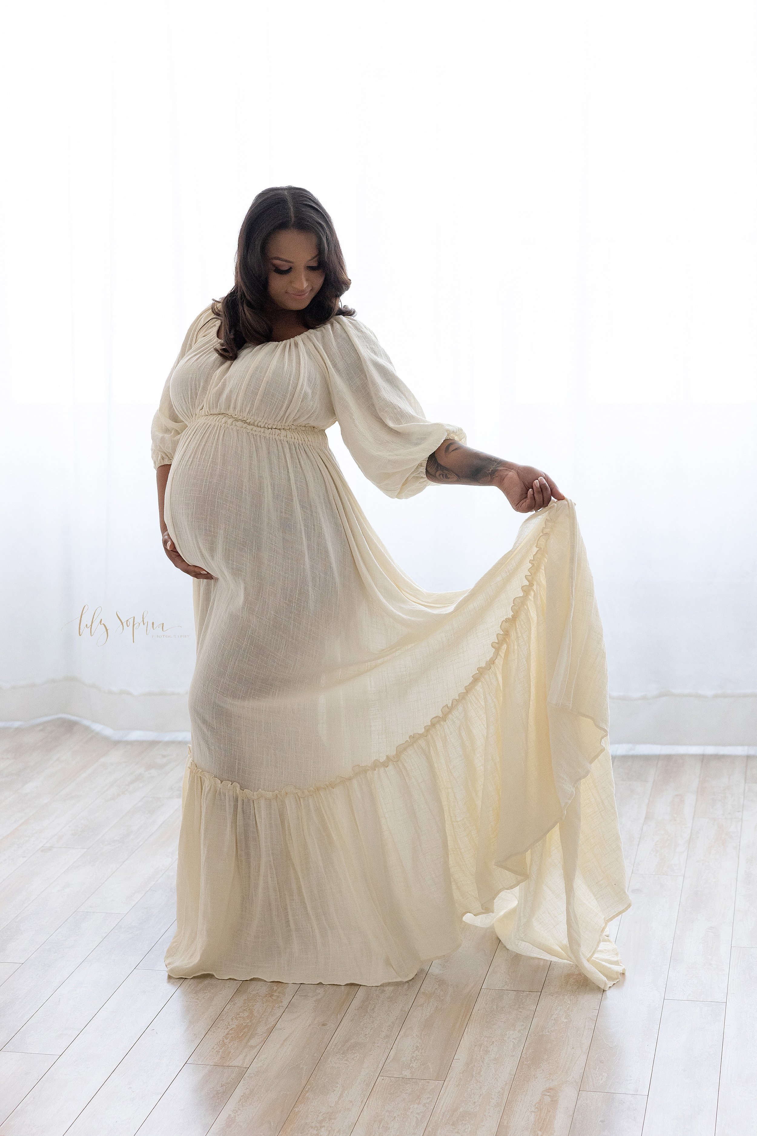  Maternity portrait of a pregnant African-American mother wearing a three-quarter sleeve peasant full-length gown as she stands in front of a window streaming natural light holding the base of her belly with her right hand and holding the ruffle of h