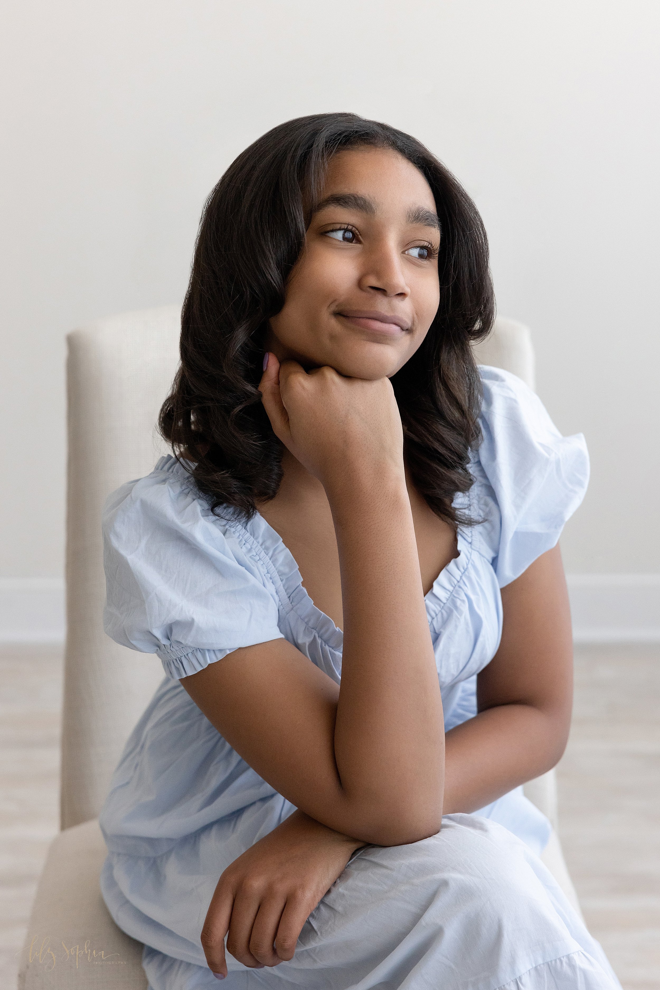  Family photo of an African-American teenager as she sits in an upholstered chair with her right hand under her chin while looking out a window streaming with natural light in a studio near Brookhaven in Atlanta. 
