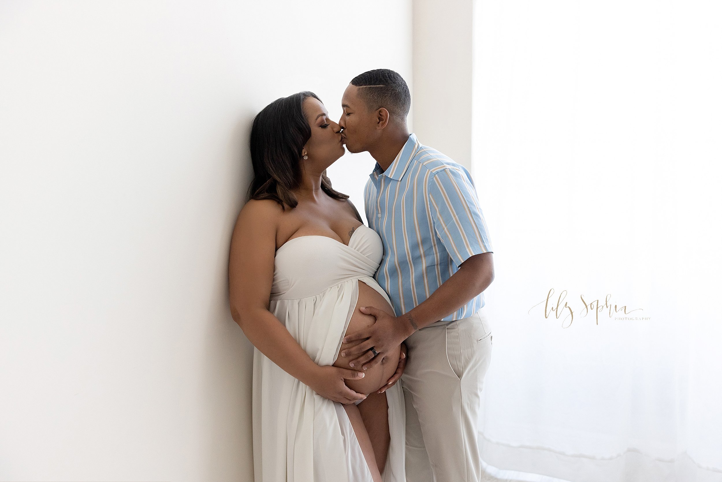 Maternity photo session of an African-American couple as the wife holds the base of her bare belly while wearing a split front gown and her husband places his right hand on their child in utero and the two of them kiss taken near Oakhurst in Atlanta