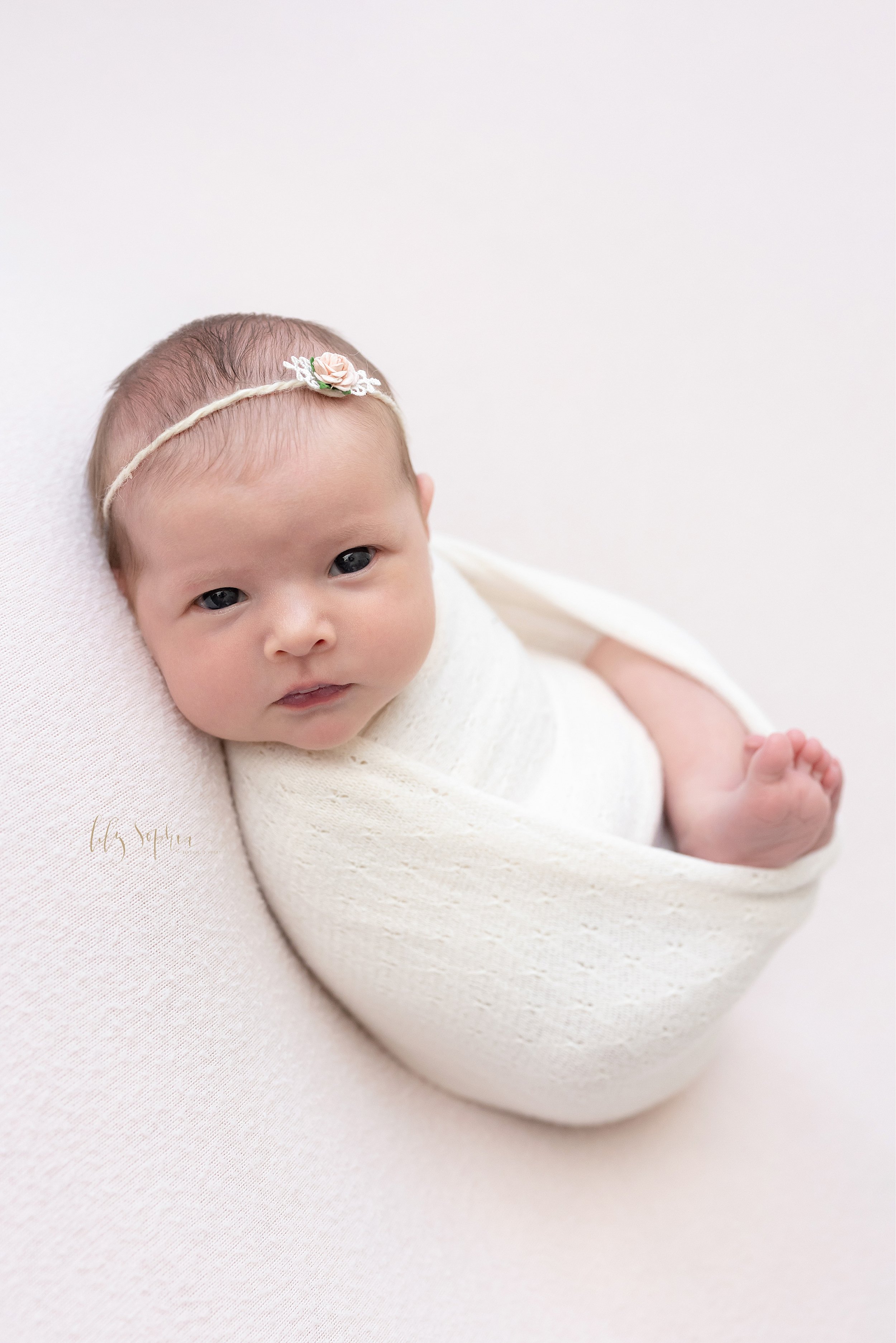  Newborn photo of an awake newborn baby girl as she is nestles in a swaddle with her right leg and foot showing as she wears a thin rose bud headband in her hair taken near Kirkwood in Atlanta in a natural light studio. 