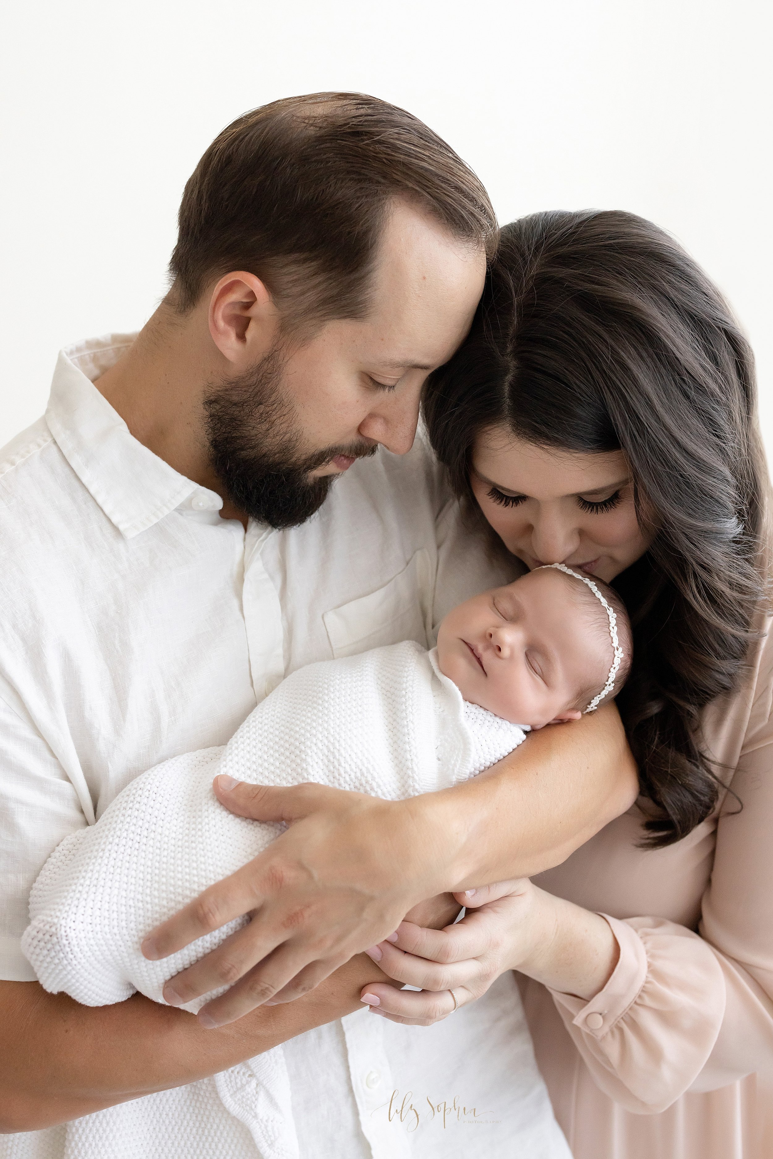 Newborn portrait of a father cradling his newborn daughter in his arms as she peacefully sleeps and his wife stands to his left side and kisses their daughter’s head taken in natural light in a photography studio near Virginia Highlands in Atlanta. 