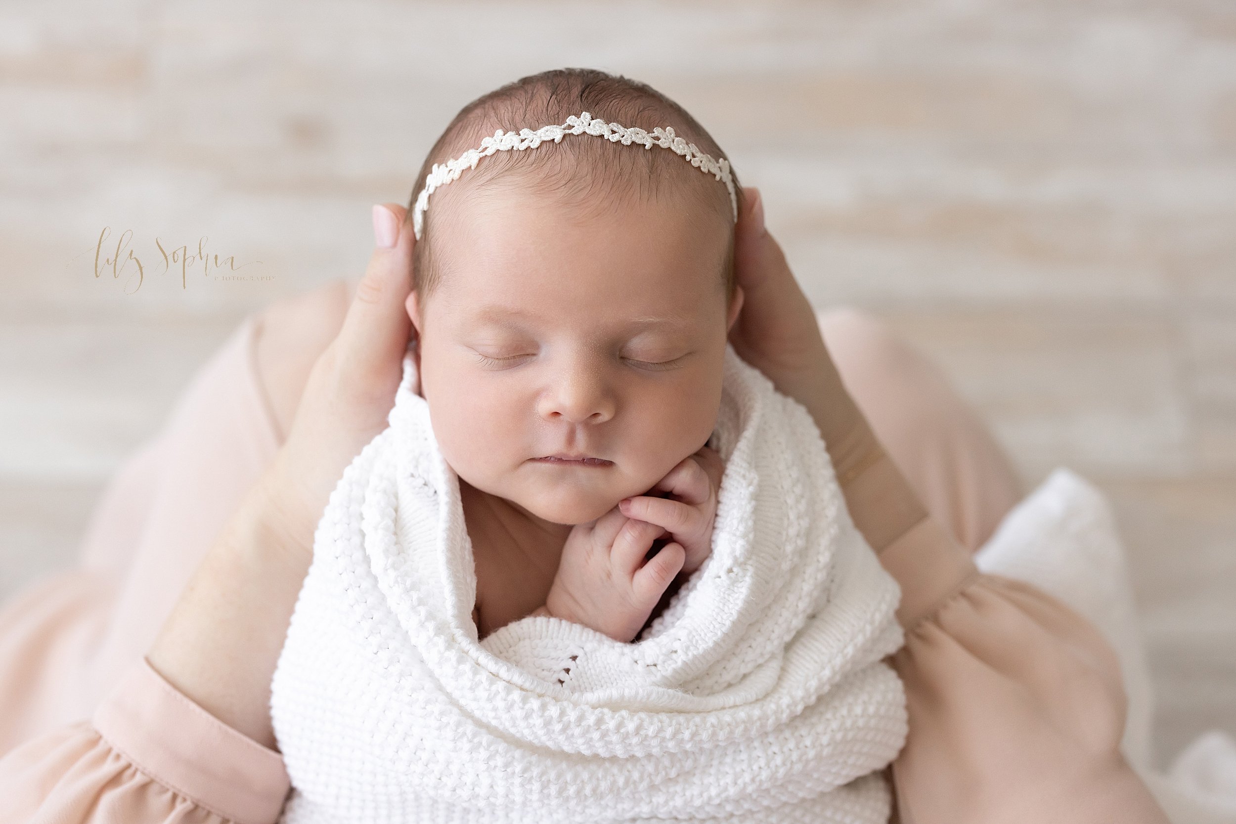  Newborn portrait of a peacefully sleeping baby girl wearing a delicate laurel headband in her hair as head is held in her mother’s hands taken in a natural light studio near Ansley Park in Atlanta. 