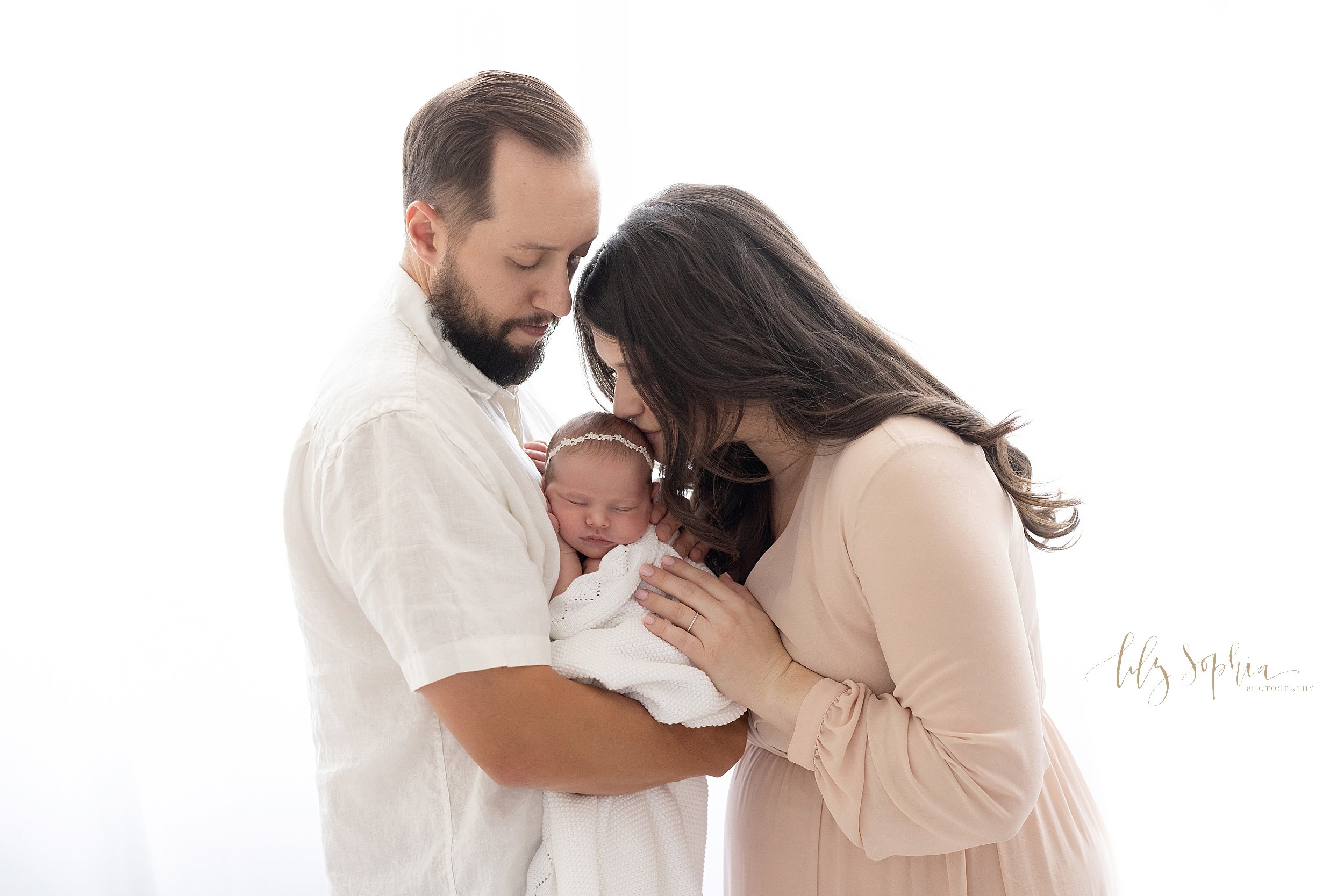  Newborn photo session with a father holding his newborn baby girl in his arms as she sleeps against his chest while mom faces dad and kisses their daughter on the crown of her head taken in front of a window streaming natural light in a studio near 