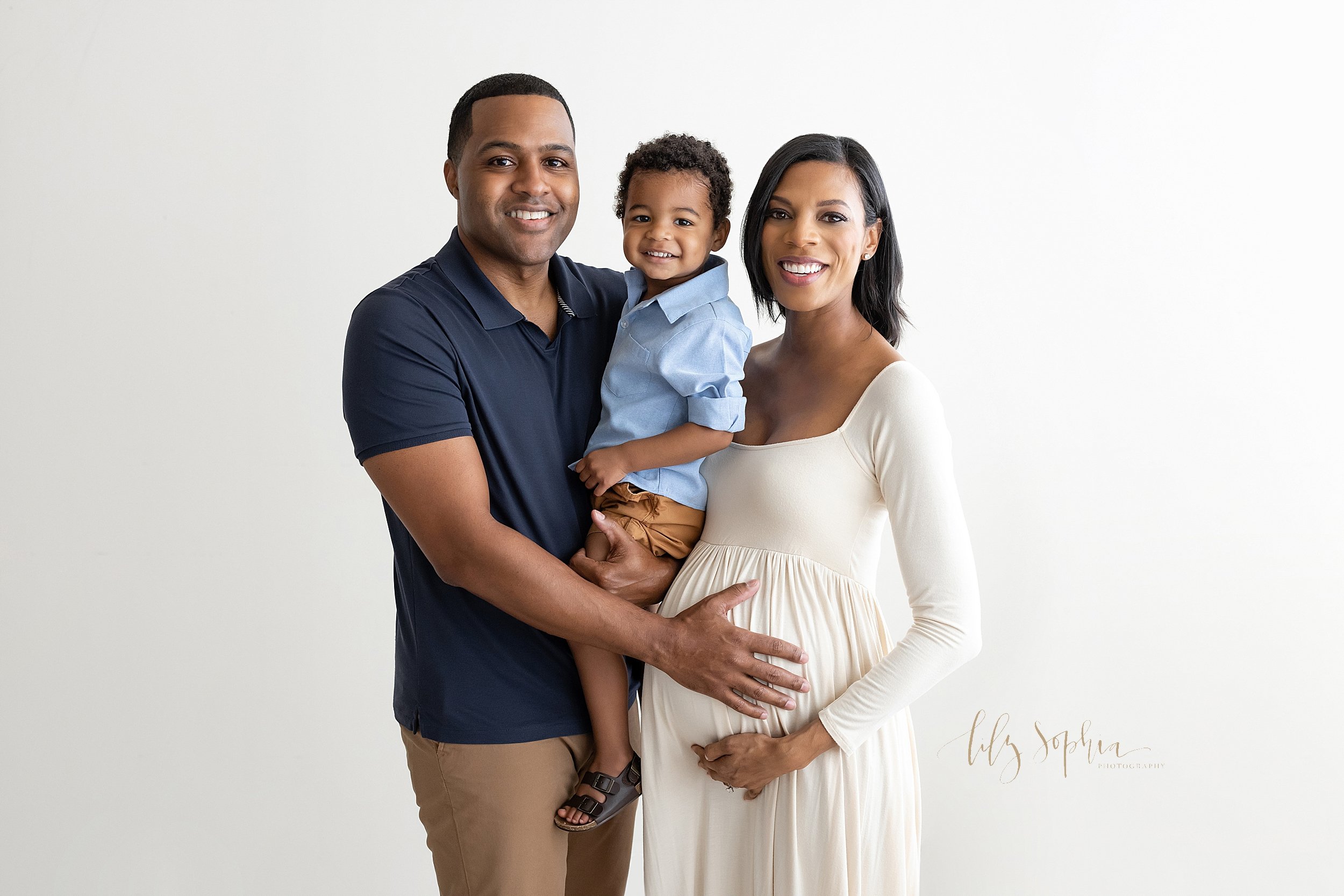  Family maternity photo of a pregnant  African-American mother standing with her right hand holding the base of her belly as she stand next to her husband who is holding their toddler son in his arms while placing his right hand on his child in utero