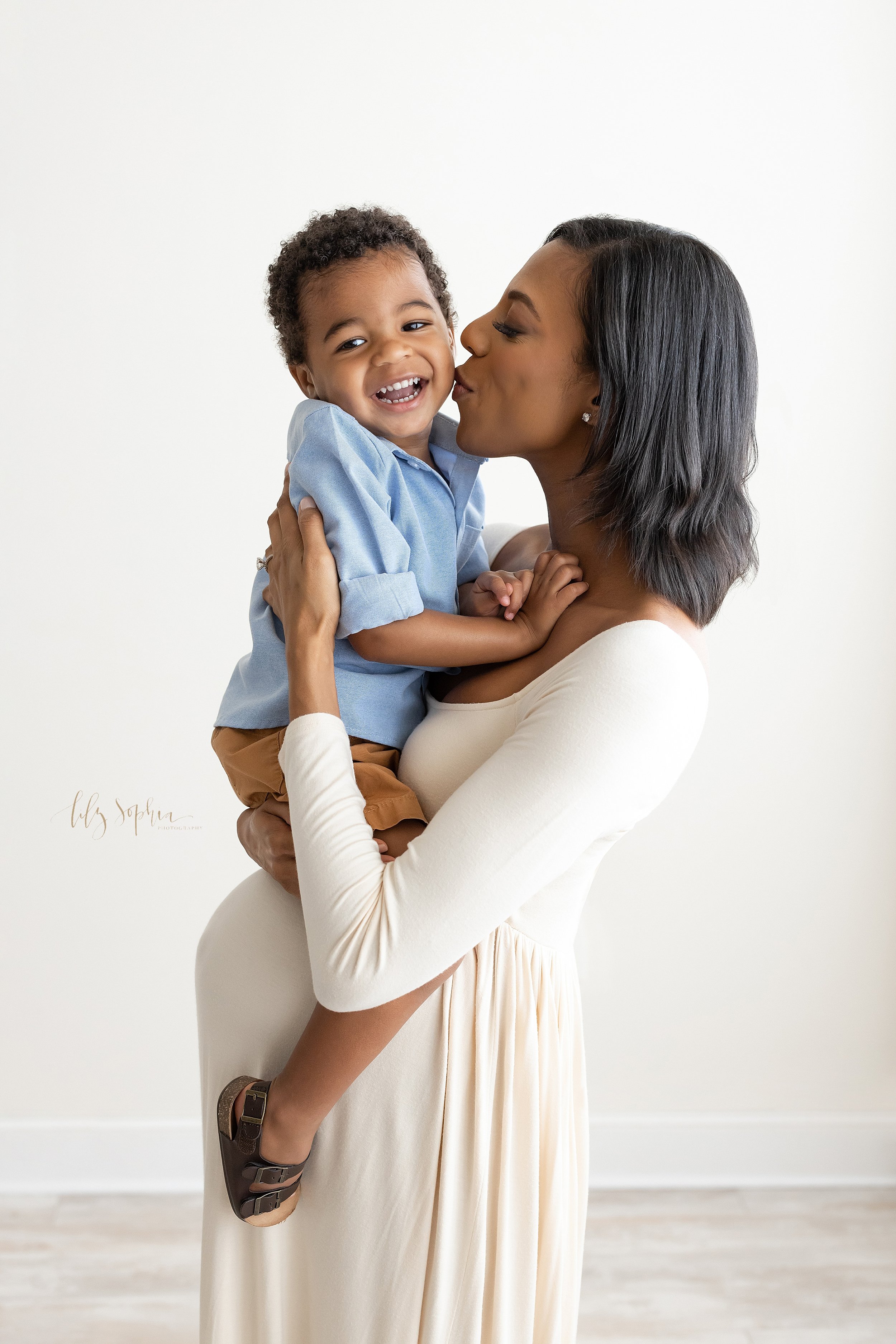  Maternity portrait of a pregnant African-American mother as she stands holding her toddler son while kissing him on his cheek taken in front of a window streaming natural light in a studio near Vinings in Atlanta, Georgia. 