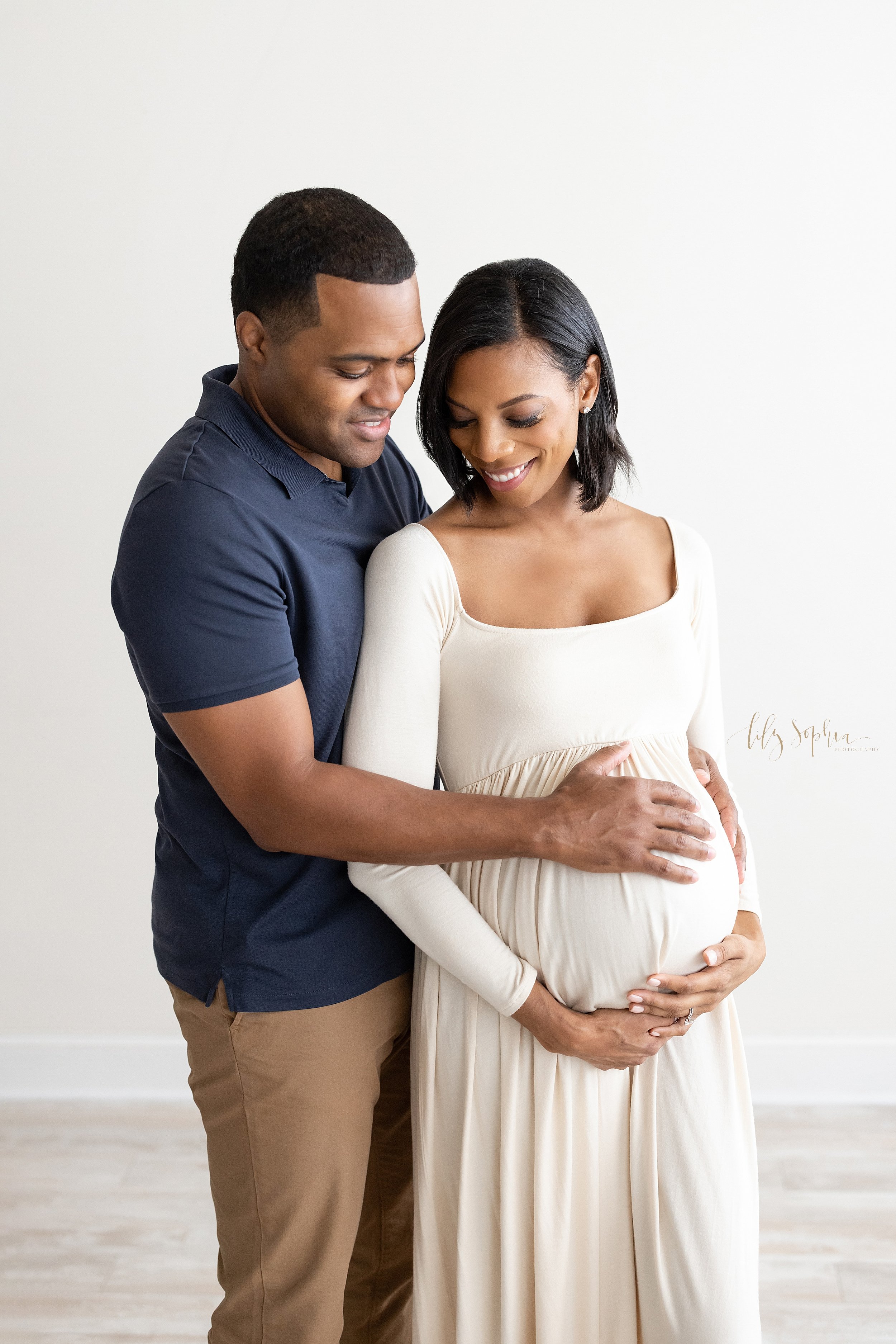  Maternity photo session with a pregnant African-American mother as she holds the base of her belly  while looking over her right shoulder and her husband reaches around her to place his hands on their child in utero taken in natural light in a photo