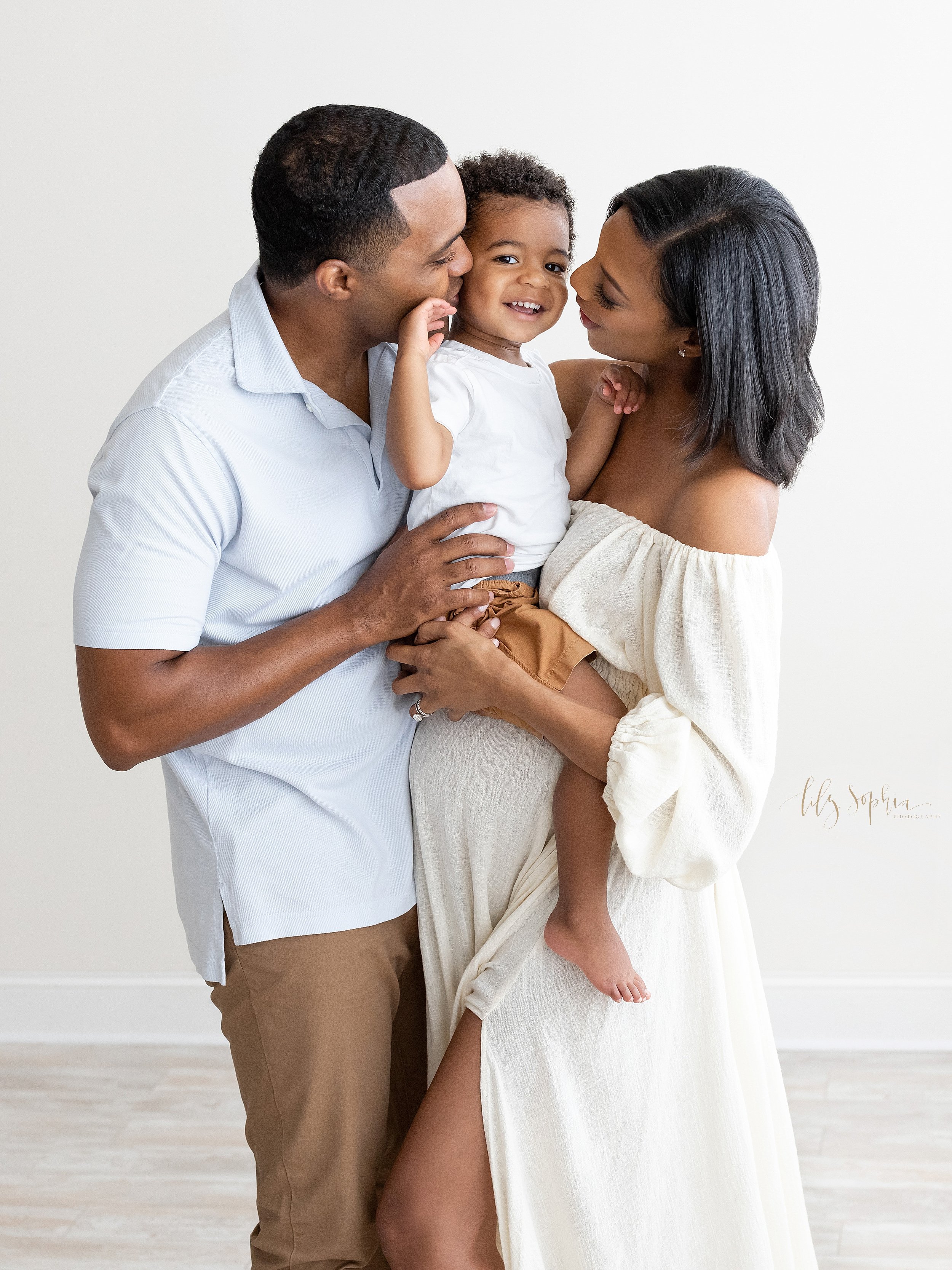  Family maternity photo shoot with the pregnant mother holding her toddler son in her arms as dad faces mom and helps to support their son with his right hand as the three of them talk together while standing in a studio near Sandy Springs in Atlanta